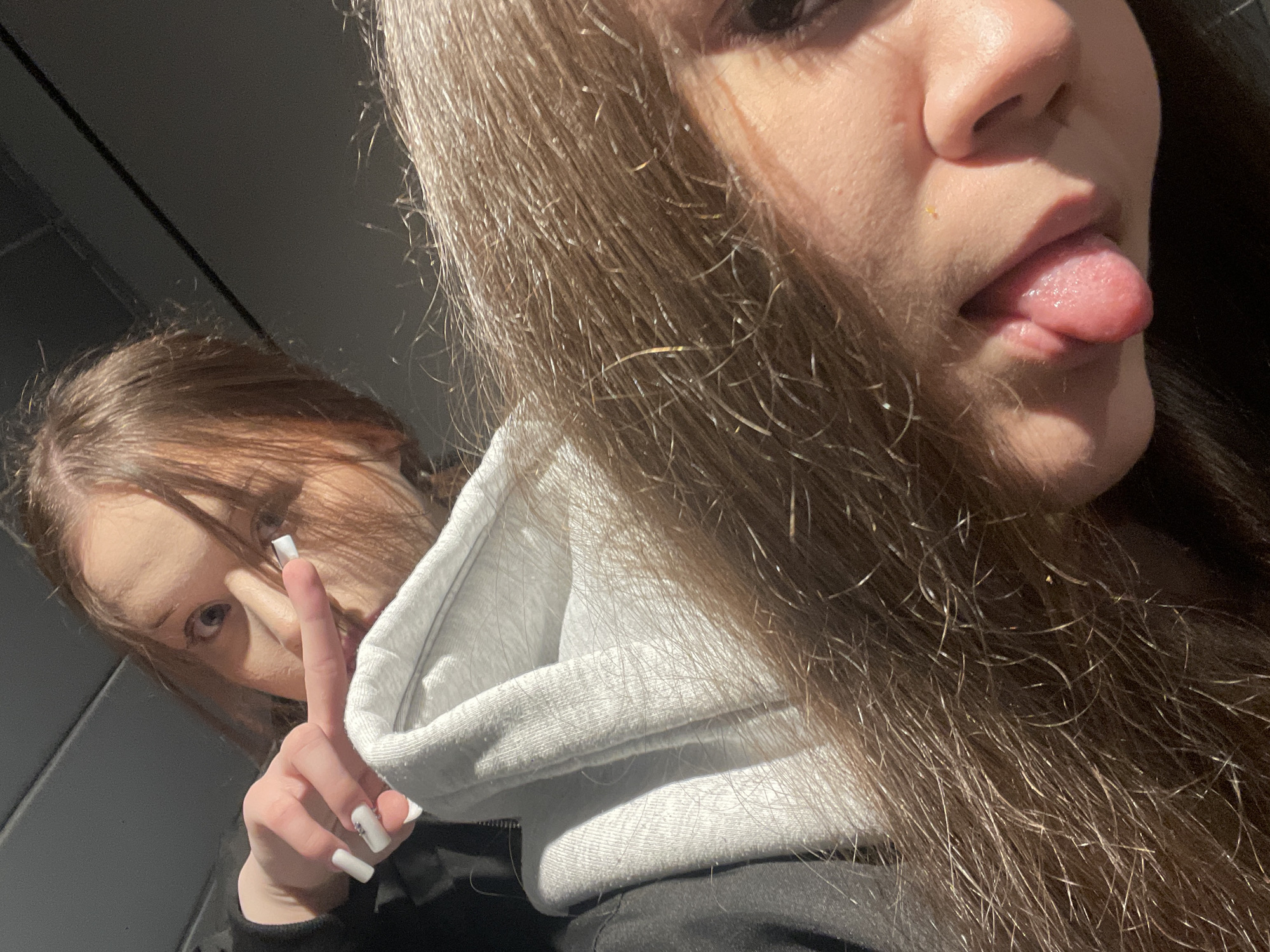 Two young women are pictured in what appears to be an indoor setting. The woman in the foreground is seen from the chest up, with her face turned to the right of the frame. She has long brown hair. Her tongue is sticking out. She's wearing a black hooded sweatshirt. The second woman is slightly behind her on the left. She is making a peace sign with her right hand, and her hand is held up near her eye. Her hair is also brown, and she has long white nails. The environment seems darkly lit. There is no indication that the location is in Chaikovsky, Russia.
