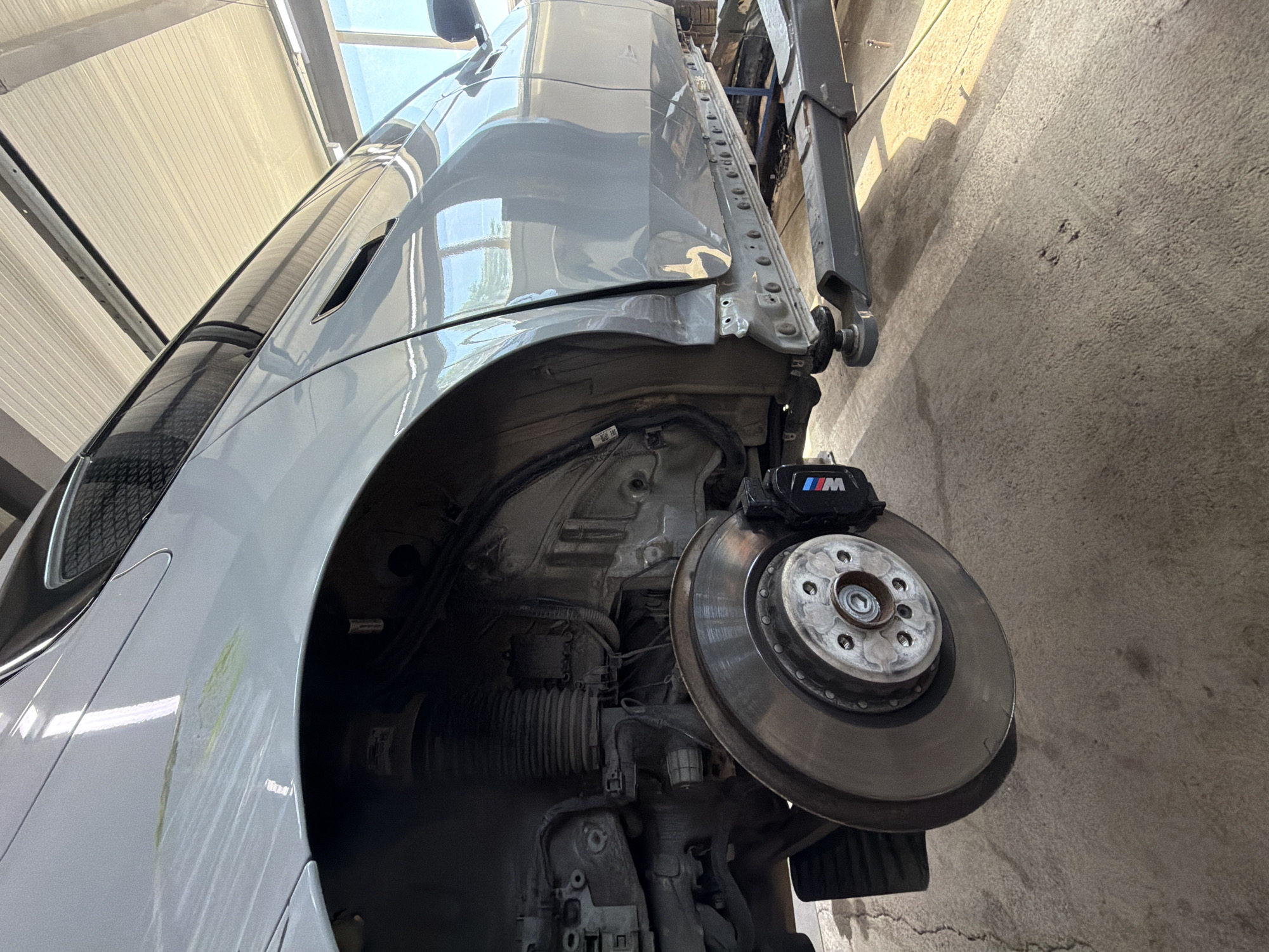 A car is being worked on in a garage. The car is gray and appears to be a BMW, as indicated by the M logo on the brake caliper. The vehicle is lifted on a hydraulic lift, exposing the undercarriage and suspension system. The focus is on the front wheel assembly, where the brake rotor and caliper are visible. The garage has a concrete floor and a corrugated metal ceiling, suggesting an industrial or workshop setting. Natural light filters in from above, creating shadows on the floor. The overall scene depicts a typical automotive repair or maintenance service. There are no people visible in the image. The time of day appears to be daytime, as suggested by the lighting. The location is identified as Burgas, Bulgaria.