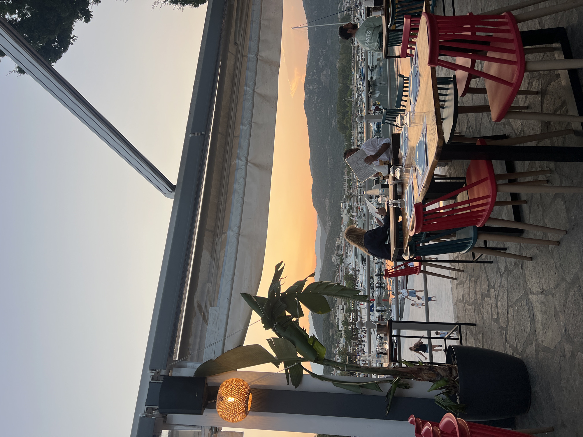A group of people are dining at an outdoor restaurant with a view of a marina and hills in the background. The sun is setting, casting an orange glow over the scene. Several tables are set with plates, glasses, and cutlery. People are seated at some tables, and others are walking in the background. The restaurant has a retractable awning overhead, and a large plant is visible in the foreground. The ground is paved with stone.
