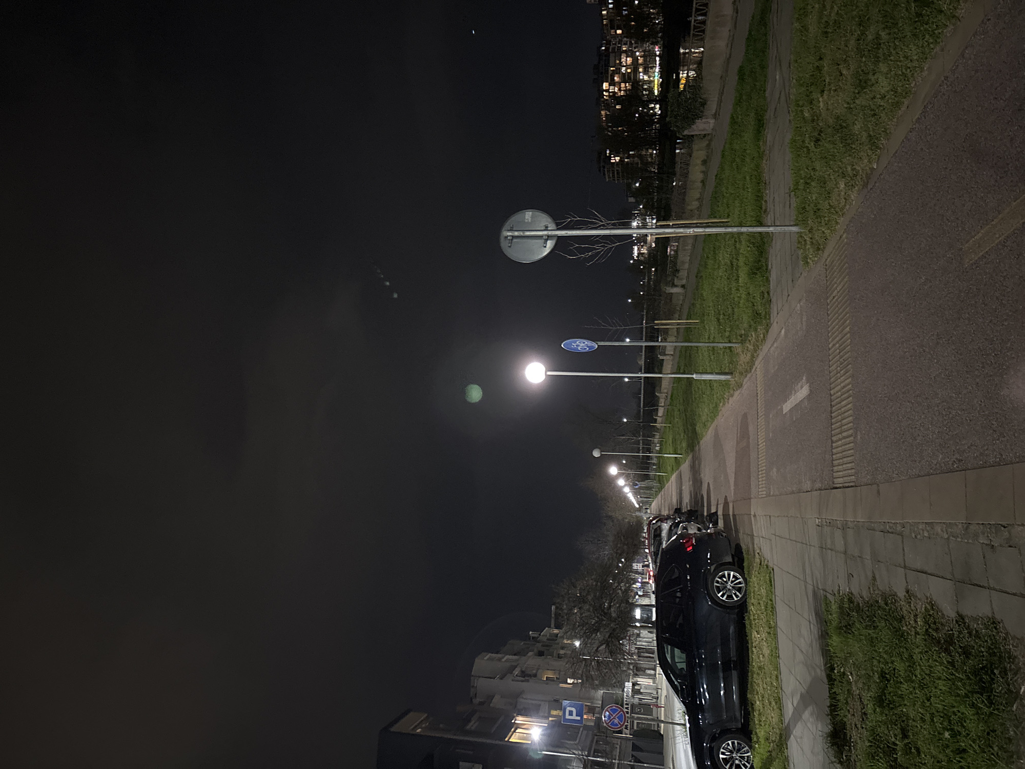 The image shows a dark, nighttime street scene, potentially in Plovdiv, Bulgaria, though that is not definitively confirmed visually. A row of streetlights illuminates the street, which has a sidewalk bordered by grass. A dark-colored car is parked along the sidewalk. Street signs and buildings are visible in the background. The sky is dark, with a few visible lights from distant buildings.