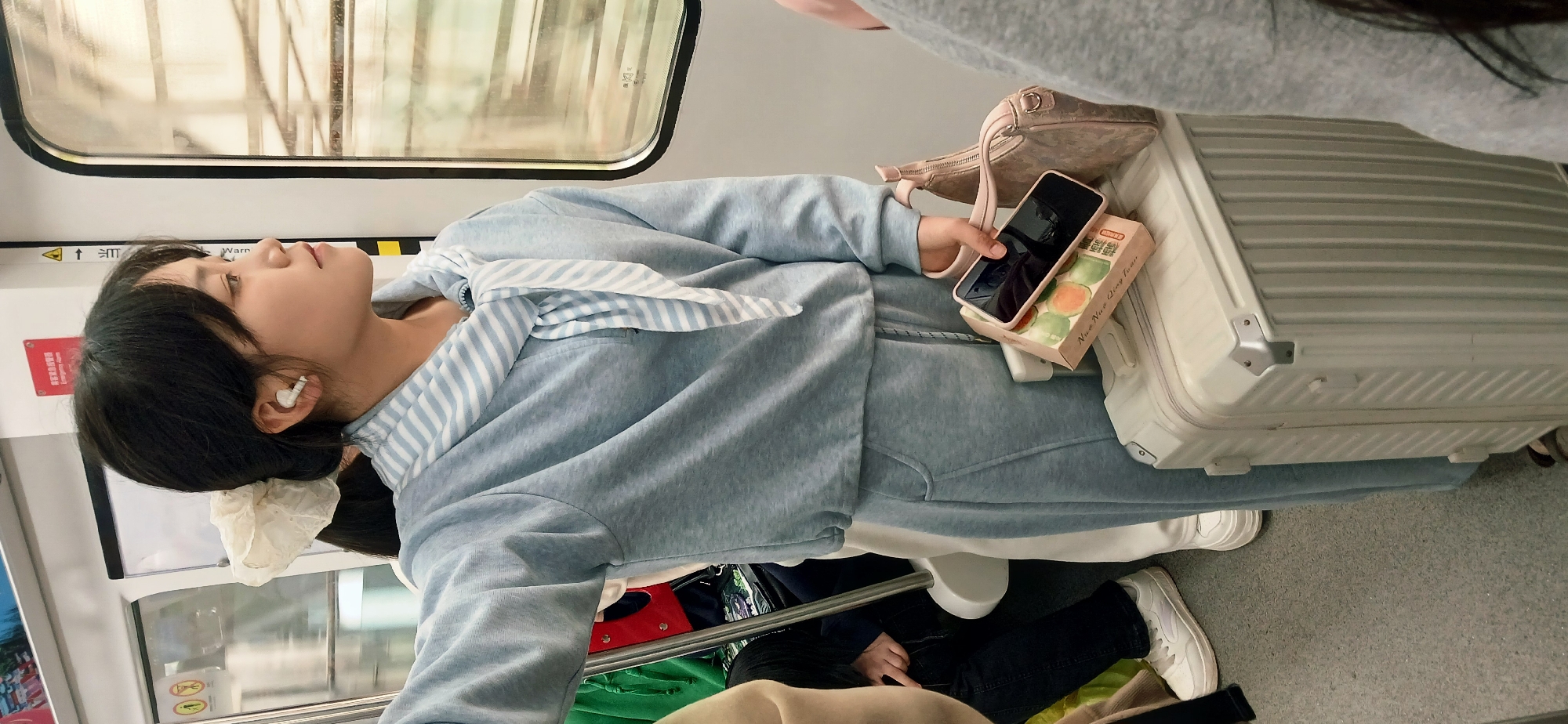 A photograph taken inside a subway car in Chongqing High-tech Zone, China, shows a young woman lying on a large, light-colored suitcase, with her head near the window. She is wearing a light blue sweat suit with a striped scarf tied around her neck and white ear buds. She is holding a black smartphone and a box of green and brown baked goods (brand "Nuo Nuo Qing Tuan") on top of the suitcase next to a light pink handbag. A second person is partially visible to the right, wearing a grey top. A second, seated person is partially visible from the knees down, wearing black pants and white sneakers. The subway car window shows reflective buildings and trees outside.