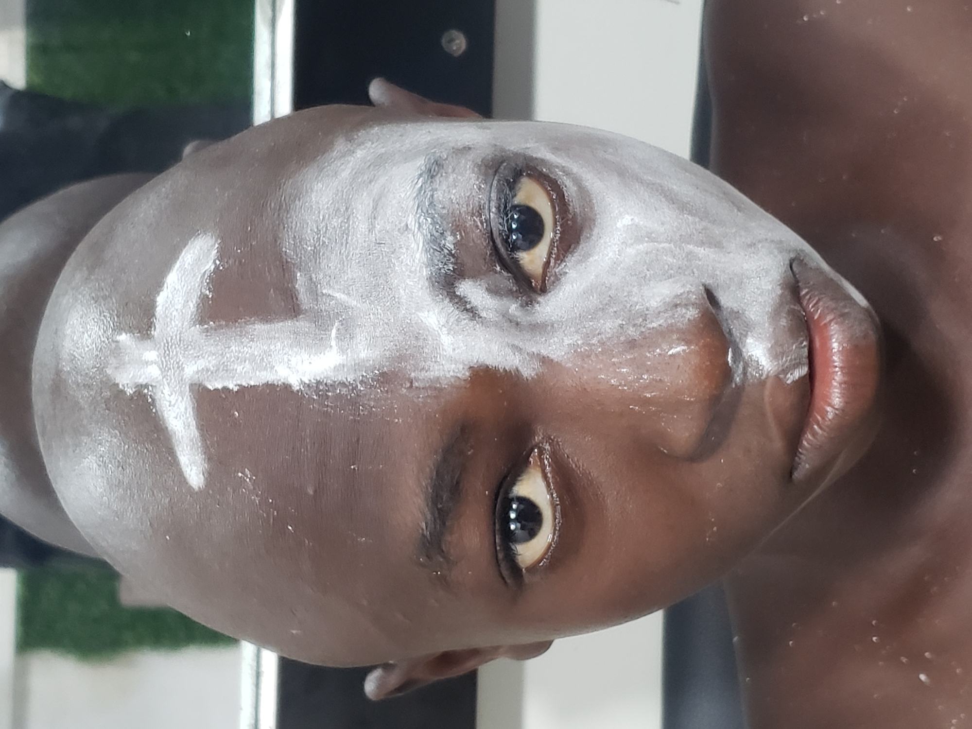 A close-up, rotated portrait captures the face of a person with dark skin, likely male, in Adankolo, Nigeria. The individual's head is tilted to the right, and they gaze directly at the viewer with a neutral expression. Their eyes are open, revealing dark irises and pupils.
The most prominent feature is the white substance applied to the person's face. A distinct white cross is visible on the forehead, stretching horizontally and vertically. The white paste or powder is also spread across the person's cheeks, nose bridge, and around the eyes, creating a striking contrast with their dark complexion. The application appears somewhat wet or oily, giving it a slightly glossy sheen in places. The person's head appears shaven or has very short hair, as no hair is visible on the scalp beneath the white markings. Their lips are slightly parted.
The background is softly blurred, showing indistinct shapes and colors, including a hint of green on the left side, possibly suggesting foliage or an artificial turf in the distance, and lighter tones on the right. The image focuses entirely on the person's face and the ceremonial or decorative markings, which may indicate participation in a cultural event, ritual, or traditional practice typical of the region. No text is visible in the image.