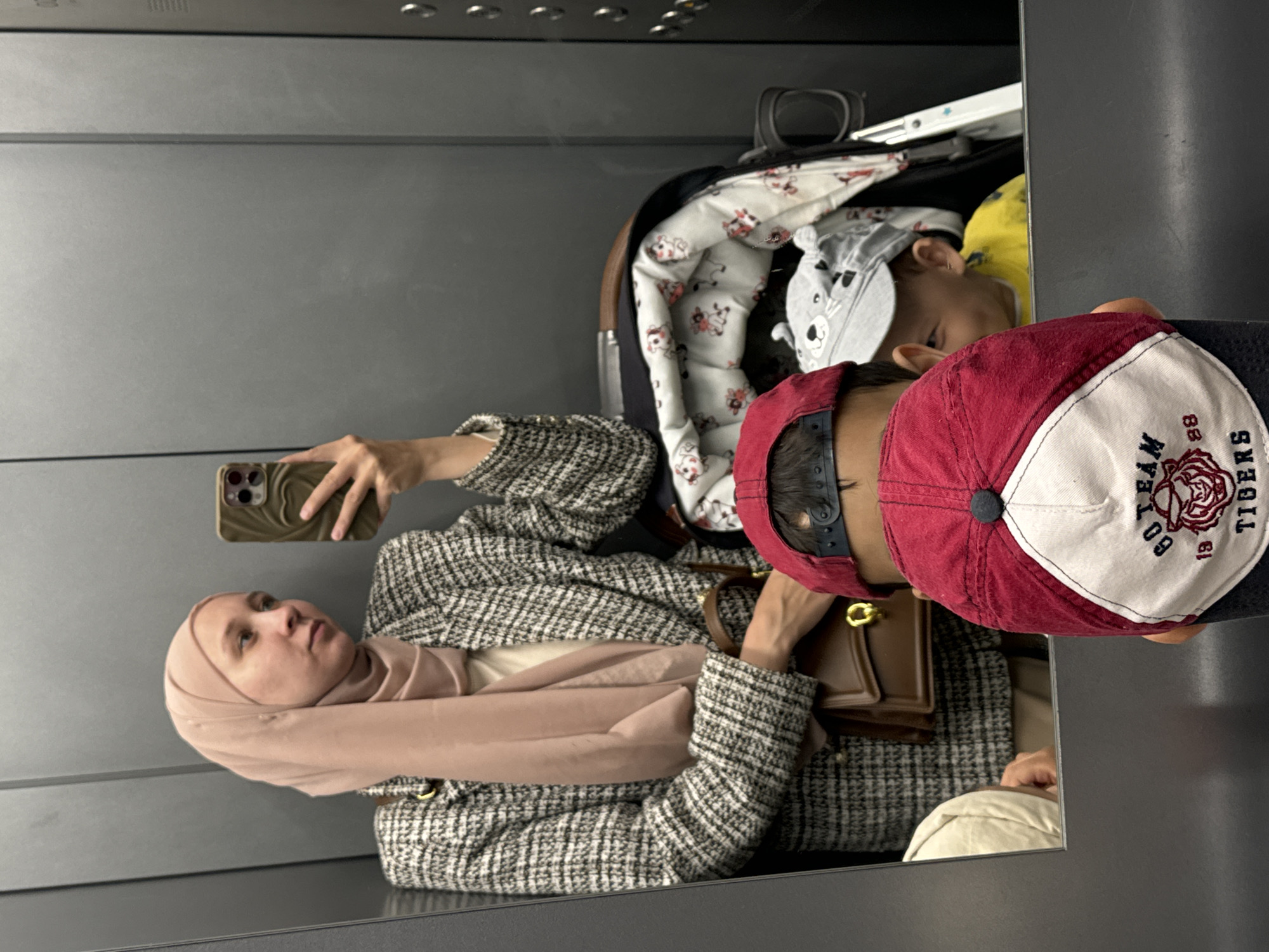 The image is a reflection of a person taking a selfie in what appears to be an elevator. The person is wearing a tweed jacket and a headscarf. A child in a baby carrier is next to them. The child is wearing a baseball cap. A second child is sleeping in the carrier. The reflection suggests the location is an enclosed space with grey walls, possibly an elevator cabin. There is no evidence suggesting that this photograph was taken in the city of Chaikovsky, Russia.