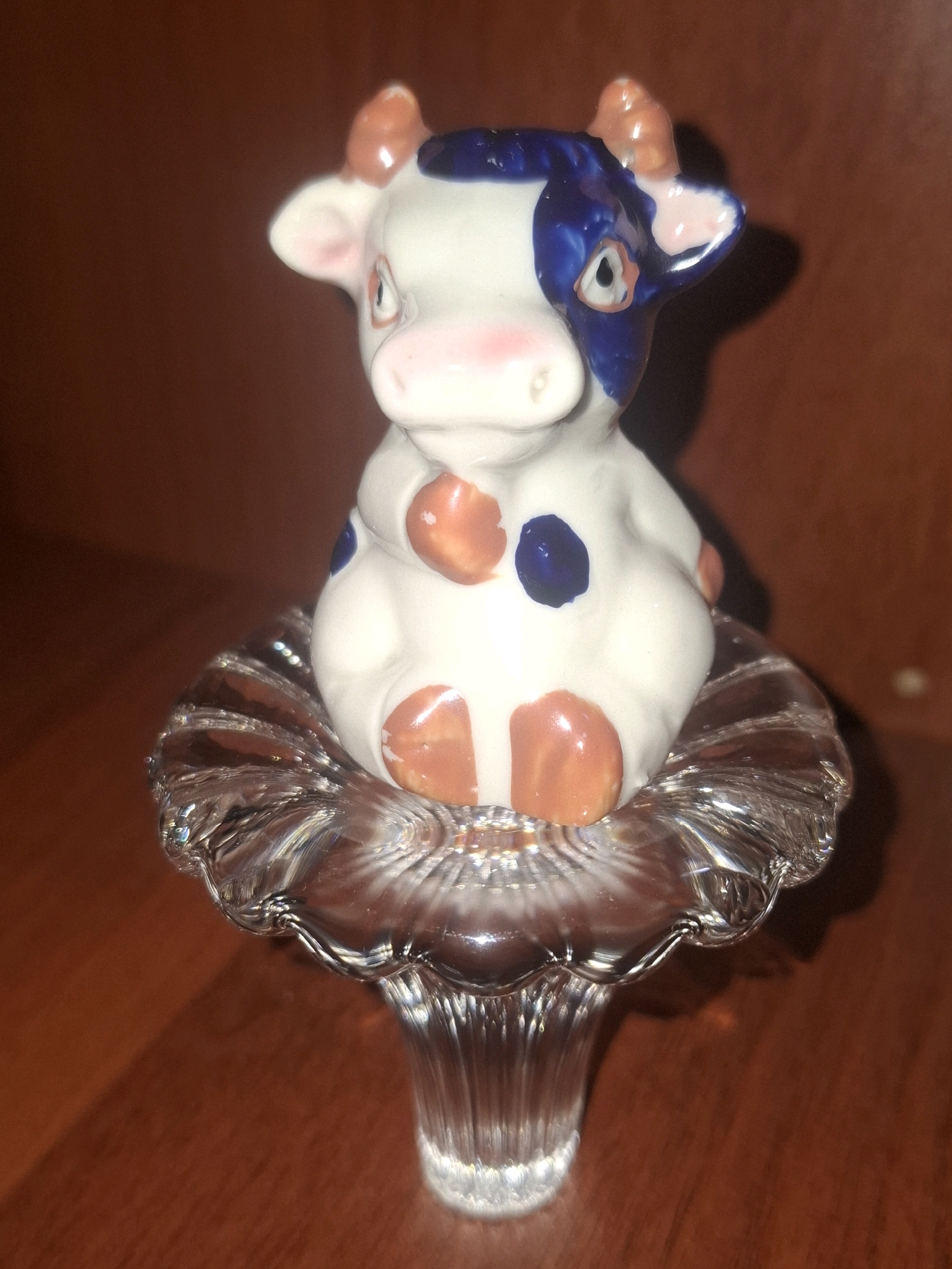 A ceramic figurine of a cow, with white, blue, and orange spots, is displayed on a clear, fluted glass stand. The cow figurine has its legs tucked underneath it, and its head is slightly tilted. The background is a warm, brown wooden surface, likely a table or shelf, with a soft shadow cast by the figurine and its stand. The lighting appears to be artificial, creating highlights on the glossy surfaces of the ceramic and glass. There is no indication of a specific location within Almaty, Kazakhstan, as the image depicts a static, indoor still life arrangement.