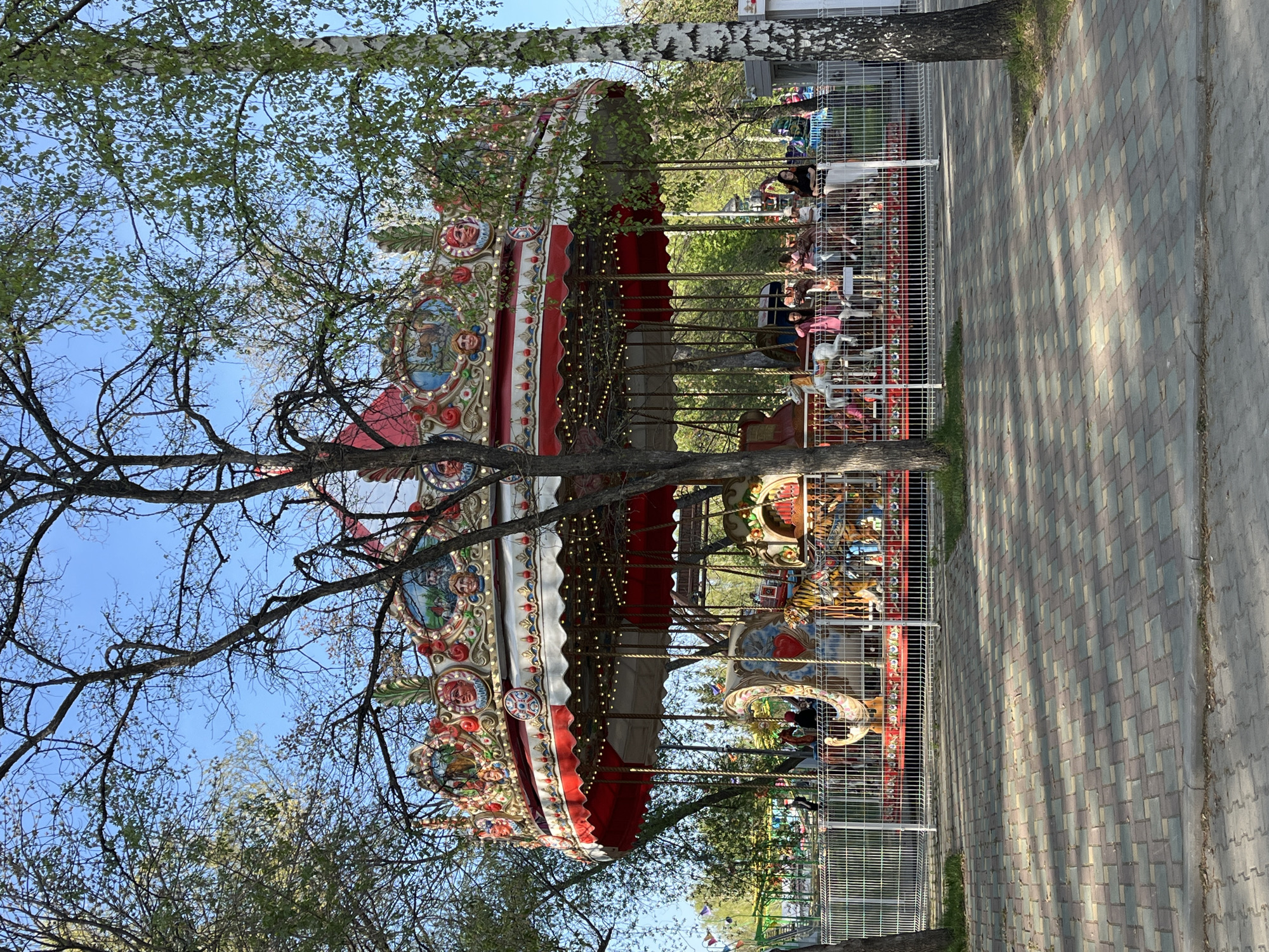 A colorful carousel with a red and white canopy is encircled by a white metal fence. People are riding the carousel. Trees are interspersed throughout the scene. The ground is a patterned brick walkway. The image depicts a carousel in the city of Yekaterinburg, Russia.