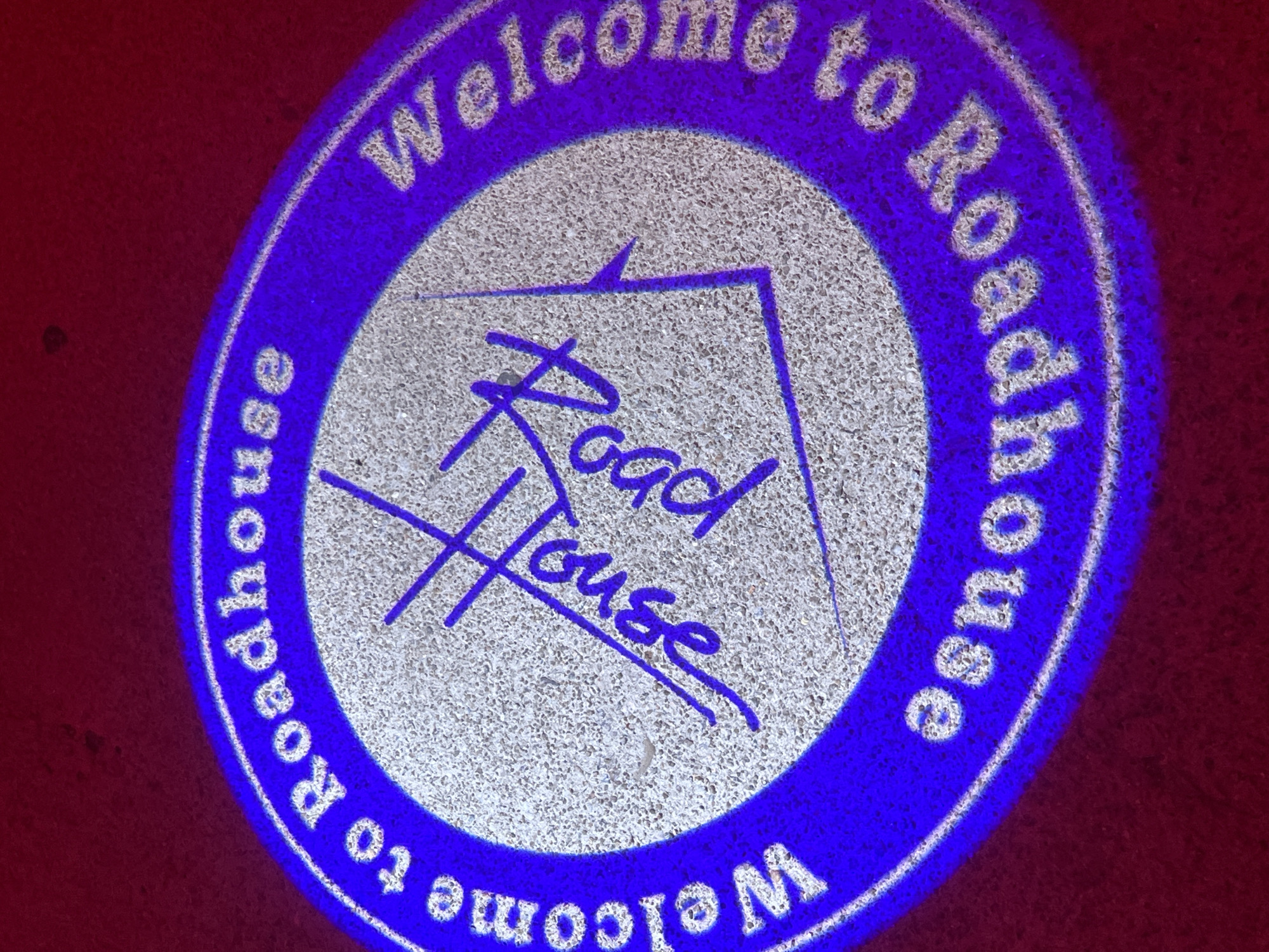 The image shows a projected logo on a surface. The logo is circular, with a blue outer ring containing the white text "Welcome to Roadhouse" repeated twice. In the center of the circle, in blue, is the text "Road House" written in a stylized script, with a crossed line motif above and below the text. The background surface is reddish-brown with a speckled texture. The location is not discernible from the image.