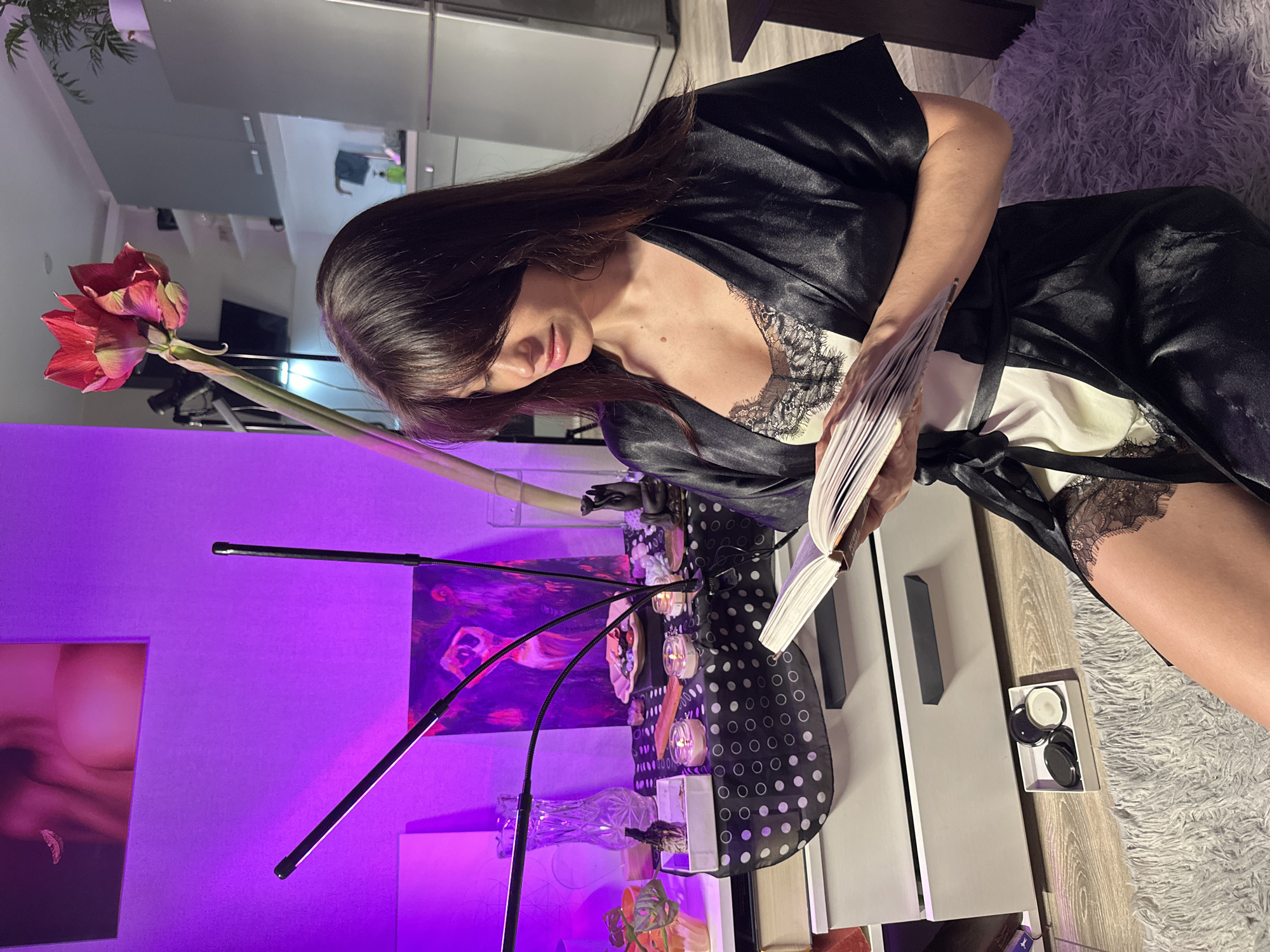 The image, taken in Murino, Russia, captures a woman engrossed in reading a book in a softly lit indoor setting, likely a living room or bedroom. She has long, dark hair and is wearing a black satin robe over a white camisole with black lace trim. She is holding an open book with both hands, her gaze directed downwards at its pages.
The room is bathed in a distinct purple ambient light, creating an intimate and serene atmosphere, suggesting it is evening or night. A tall, elegant red amaryllis flower with multiple blooms stands prominently in the foreground to the left. Near the flower, two flexible black LED lights extend, casting additional illumination.
A table or shelf is visible to the woman's left, partially covered with a black and white polka dot patterned fabric. On this surface, various decorative items are arranged, including small, illuminated objects resembling candles or lights, glass vases, and possibly small plants or other ornaments. Two art prints or paintings hang on the purple wall behind the table. A soft, white, shaggy rug covers part of the floor, and a white box containing several black circular lids is placed on a light-colored surface near the rug. No discernible text is visible on the book or other objects in the frame.