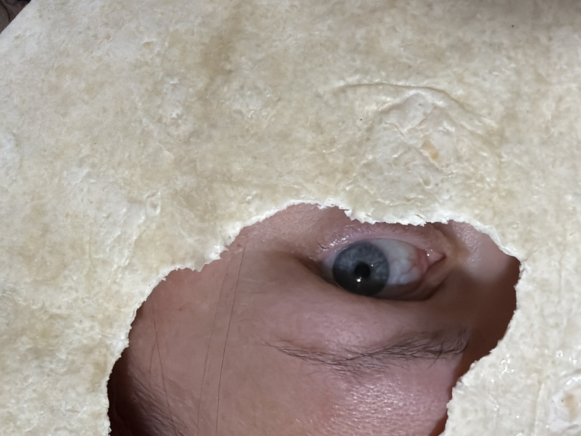 A close-up shot depicts a person's face partially obscured by a pale, textured object with a jagged hole. The person's left eye, blue with visible blood vessels, is visible through the opening. Skin and a few facial hairs are also visible. The textured object appears to be a thin, pliable material, possibly a tortilla or similar flatbread. There is no indication of the location being in Chaikovsky, Russia.