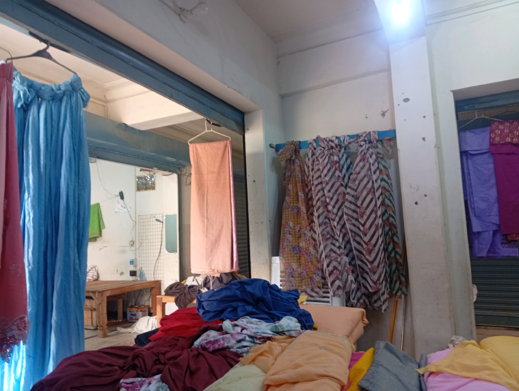 A shop interior in Larkana, Pakistan, is filled with hanging and piled textiles. In the foreground, a pile of colorful fabrics, including shades of burgundy, blue, peach, and yellow, are stacked on top of each other. Behind this, a wooden table sits in the center of the room, with more fabrics on top and around it. To the left, several large pieces of fabric, including a bright blue one, hang from hangers. The shop appears to be a textile or clothing store, with rolled fabrics and various garment pieces visible. The walls are painted white, and a fluorescent light fixture is visible on the ceiling. A metal roller shutter is partially visible in the background, suggesting it is a commercial establishment. The overall impression is one of a busy, possibly cluttered, retail space for fabrics.