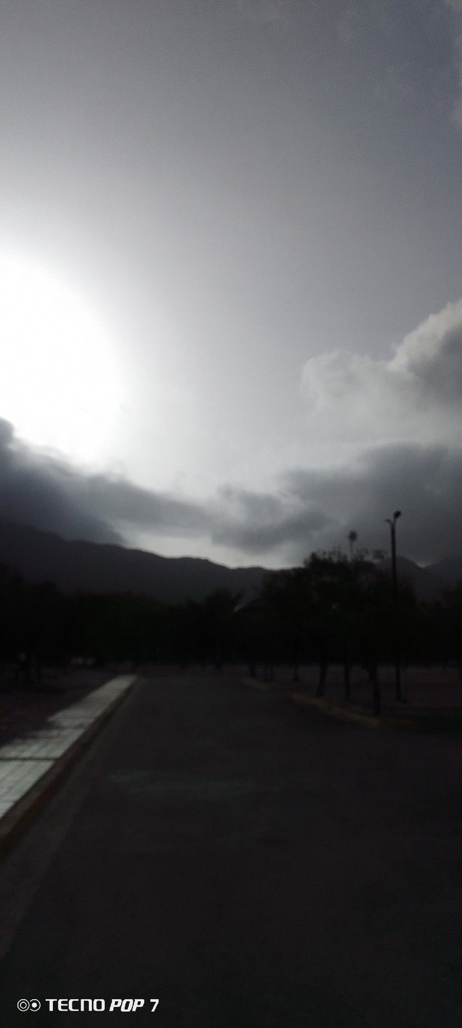 The photograph depicts an outdoor scene in Pedro González, Venezuela under an overcast sky. The sun is partially obscured by thick clouds, creating a diffused, high-contrast lighting condition.
In the middle ground, a silhouette of mountains is visible against the sky. In the foreground, a dark paved road stretches into the distance with a sidewalk visible on the left. Trees are present along the road's edge, appearing as dark outlines against the lighter sky. A single lamppost is visible among the trees.
The scene is generally devoid of people, and no specific actions or interactions are discernible. The overall environment suggests a park or public space adjacent to the road.