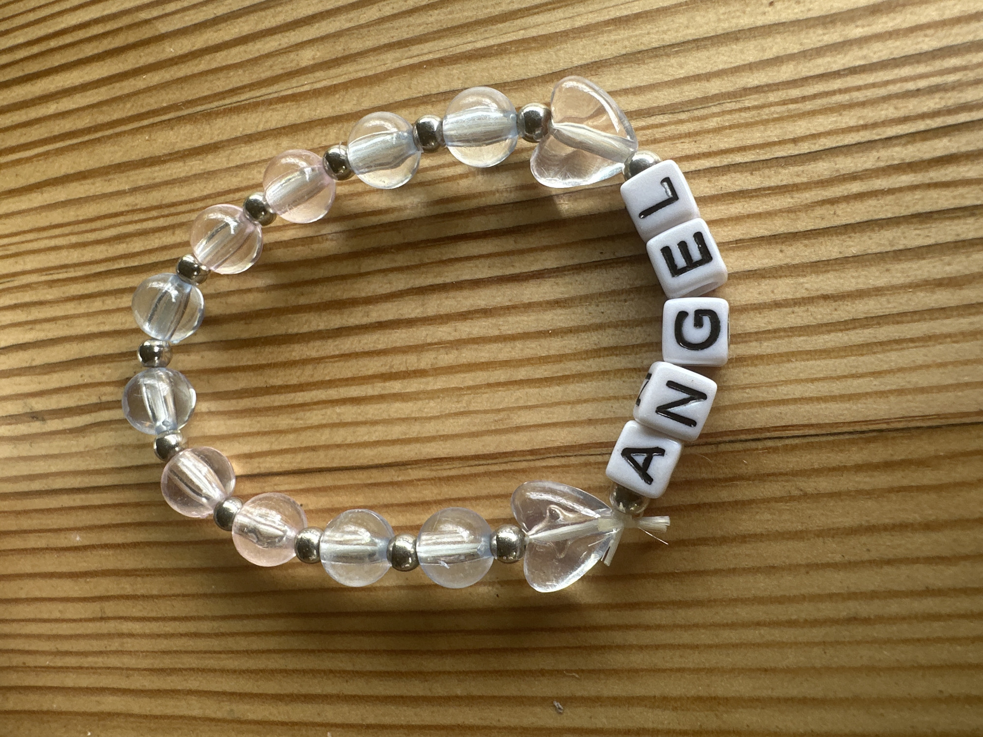 A beaded bracelet with the word "ANGEL" spelled out in white letter beads lies on a wooden surface. The bracelet is composed of clear and pinkish-clear faceted beads, interspersed with silver spacer beads. Two clear heart-shaped charms are also incorporated into the bracelet. The wooden surface appears to be a table or countertop with prominent grain lines. There are no people visible in the image, and no specific location in Almaty, Kazakhstan, can be identified from the provided visual information. The scene is static and depicts a single object.