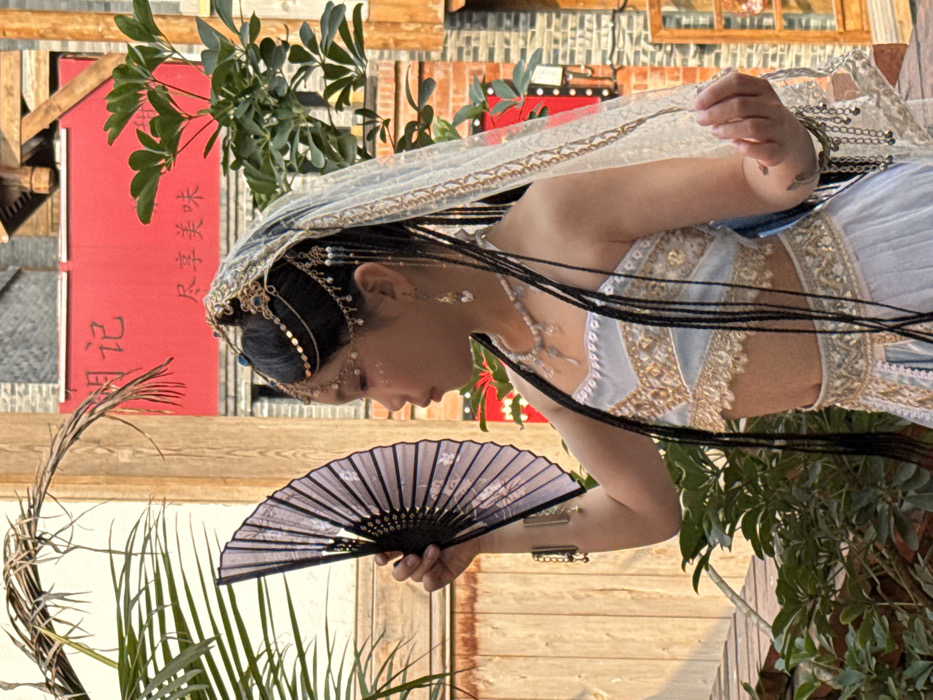 A person in an elaborate, ornate costume holds a fan. The costume is light blue and gold, with intricate detailing. The person also wears a decorated veil and headpiece. The background features a wooden structure and greenery.