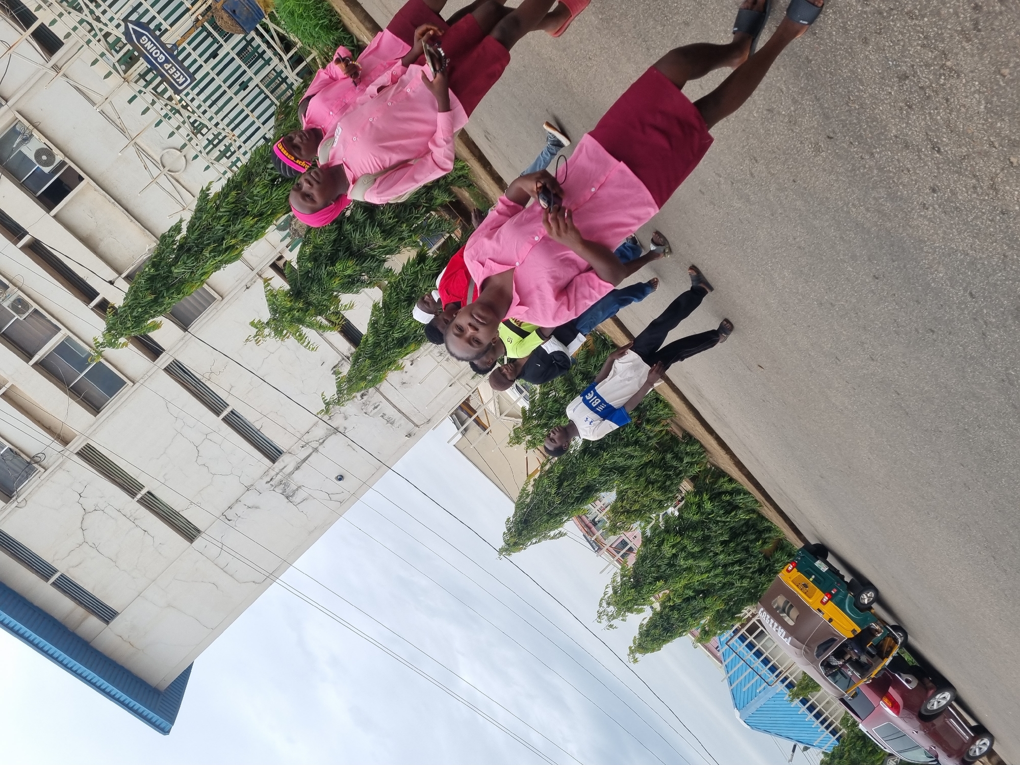 The image depicts a street scene in Jos, Nigeria. A group of individuals are walking along the roadside. Three women in the front, who are wearing pink shirts and dark red skirts, seem to be looking at their phones. Behind them are several other individuals in casual clothing. A multi-story building with light-colored facade and multiple windows is visible on one side of the street, partially obscured by green vegetation. On the other side of the street, there are parked cars, including a red sedan and a taxi. A sign is visible on the building. The sky is overcast.