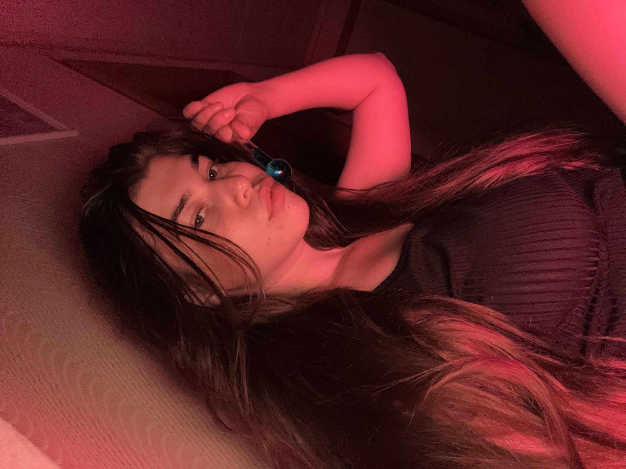 The image depicts a young woman with long, dark hair, holding a small glass vial filled with blue liquid up to her face. She is wearing a dark, ribbed top. The lighting in the scene is predominantly red, casting a strong red hue over her skin and hair, with some areas appearing darker. The background is dimly lit and indistinct, showing textures that could be wallpaper or fabric. There is no visible indication of the location being in the city of Мещёры, Russia, as this is a self-portrait with no discernible environmental context.