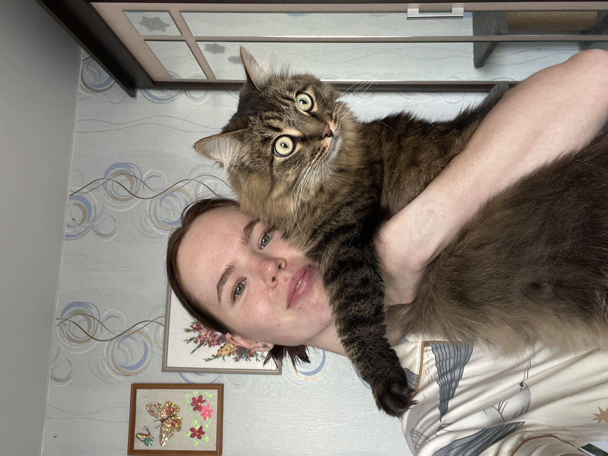 A woman with fair skin, light blue eyes, and light brown hair is holding a large, fluffy brown and black cat with green eyes. She is looking directly at the camera with a neutral expression. The cat is also looking at the camera with a slightly quizzical expression. The woman is wearing a patterned pajama top. The background includes a patterned wallpaper with light blue and brown swirls. There are two framed pictures on the wall. The upper frame depicts a floral arrangement and the lower frame contains a butterfly with flowers. A dark wooden trim is visible at the top of the frame, suggesting a cabinet or door frame. The scene is located in Chaikovsky, Russia.