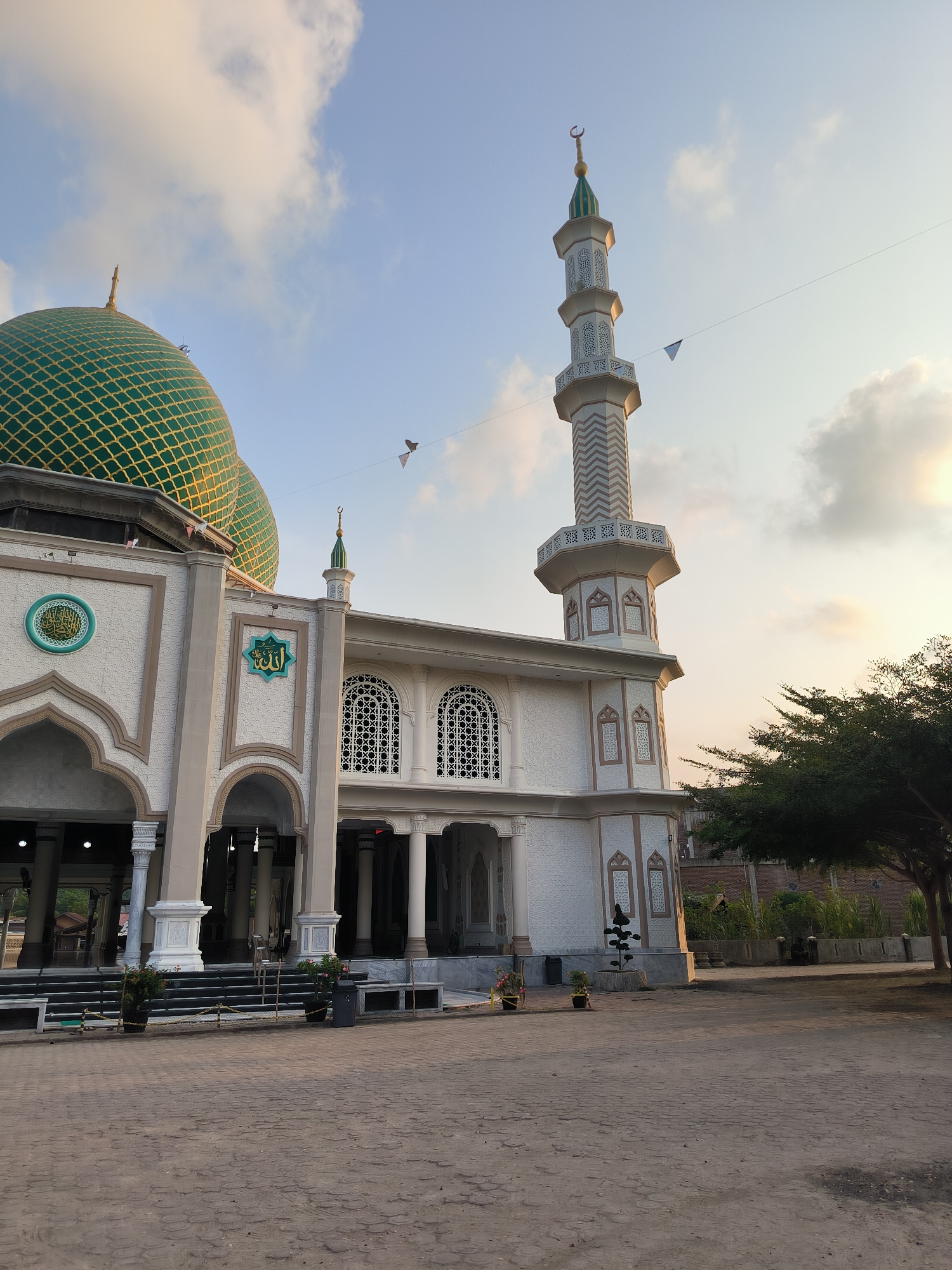 A mosque, likely the Islamic Center Lhokseumawe, is pictured under a partly cloudy sky at what appears to be sunset. The mosque features a large green dome with a gold latticework pattern and a tall, slender minaret topped with a crescent moon. The facade of the mosque is white with light brown accents, adorned with decorative Islamic calligraphy. To the right of the mosque, a lush green tree with a thick trunk is visible, and behind it, a brick wall and some vegetation can be seen. The foreground is a paved courtyard with a textured, brick-like pattern. There are no people visible in the image.