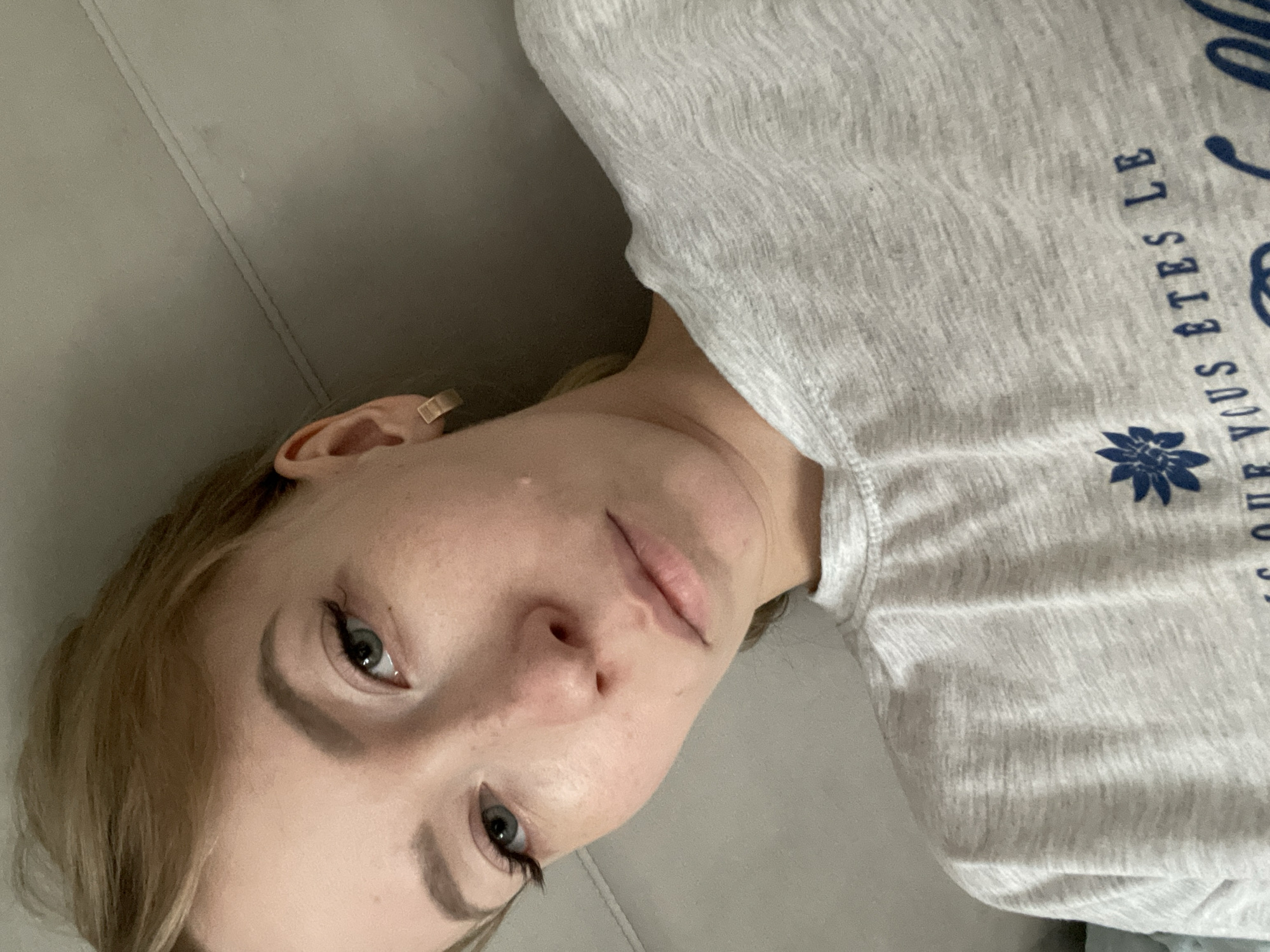 A woman with light blonde hair and blue eyes is lying on a light gray surface, possibly a couch or bed. She is wearing a light gray t-shirt with blue lettering and a flower design. The text on the shirt appears to be in French. She is facing the camera with a neutral expression. A piece of gold jewelry is visible in her left ear. There is no discernible information about the location being Perm, Russia.