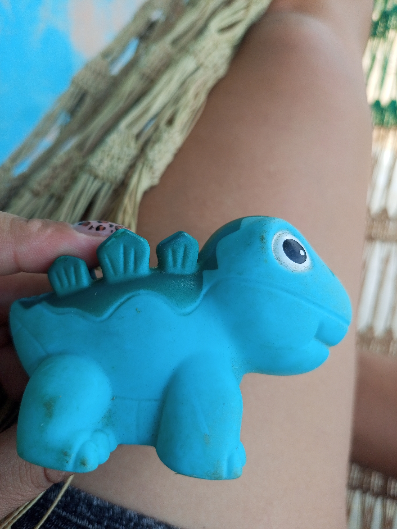 A close-up, eye-level shot shows a person's hand holding a turquoise dinosaur toy. The toy has green accents on its back and a white and black eye. The background consists of a person's legs, partially covered by a green woven blanket, and a hint of blue sky. The image is undated and its location in La Tagua, Venezuela, is unknown.
