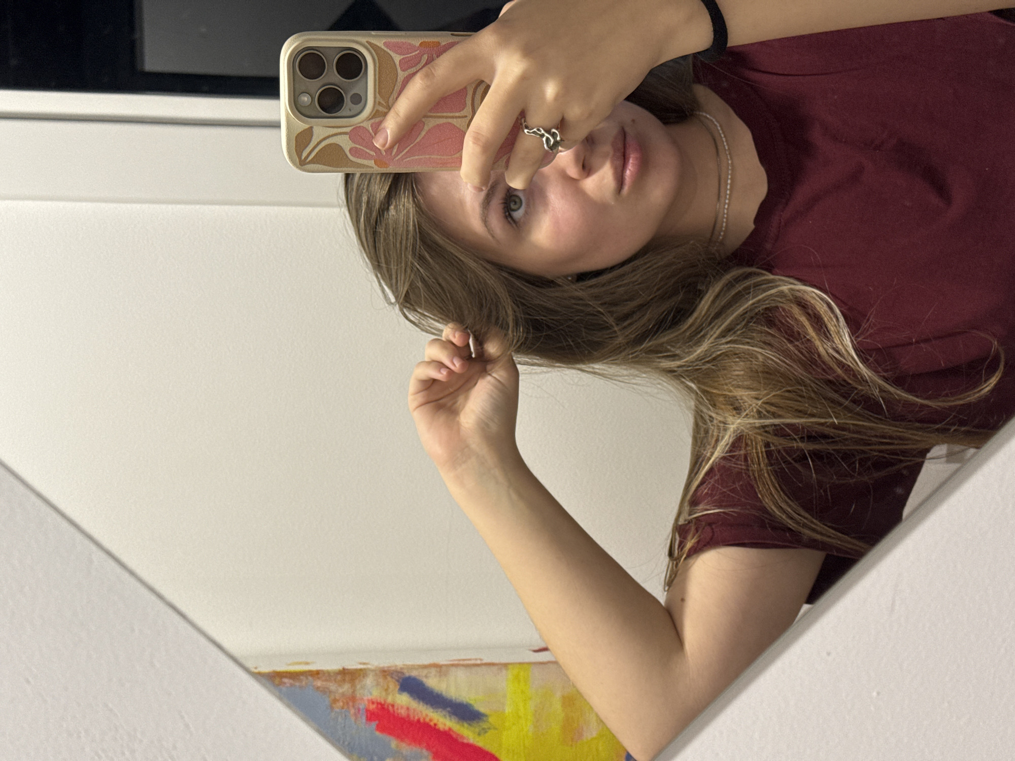 A young woman with long, highlighted blonde hair is taking a selfie in a mirror. She is wearing a maroon t-shirt and a silver necklace. Her phone has a floral-patterned case, and she is wearing a silver ring. A painting with red, blue, and yellow paint is partially visible in the mirror's reflection. The background includes a white wall and a window with black trim. This image was taken in an indoor setting, but there is no information on the city of Vozdovac Urban Municipality, Serbia.