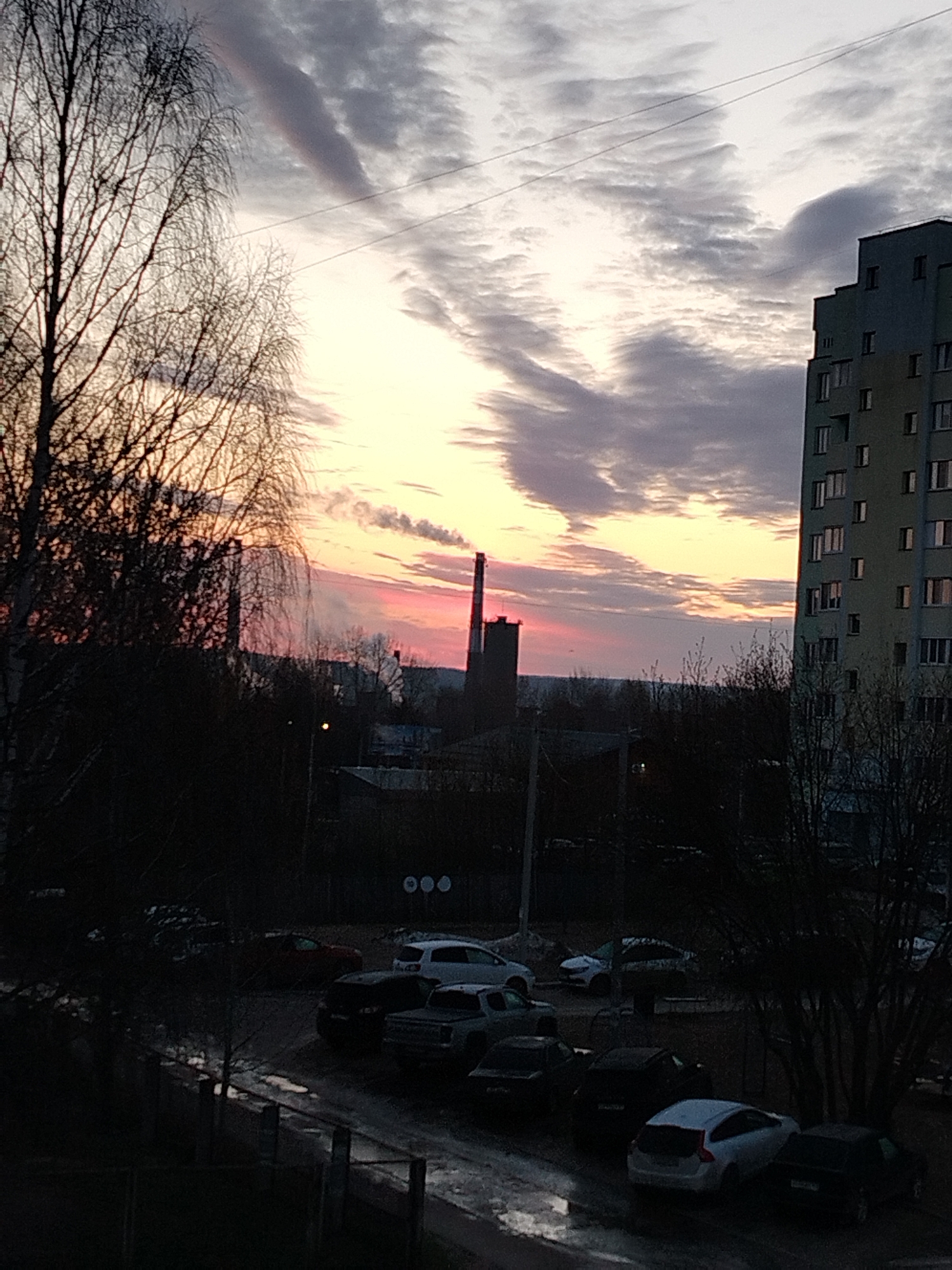 The image depicts a city scene at dusk or dawn, in Unknown, Unknown. The sky is filled with dramatic clouds tinged with pink and yellow from the rising or setting sun. A tall building is visible on the right side of the frame, characterized by numerous windows arranged in a grid-like pattern. In the midground, a smokestack rises above the surrounding structures, emitting a faint plume of smoke. The foreground features several parked cars on a wet or icy surface, likely a parking lot or street. Bare trees frame the image on the left and right, suggesting a late fall or winter season. Overall, the scene presents an urban landscape with a focus on the interplay between nature and industry, under a dynamic sky.