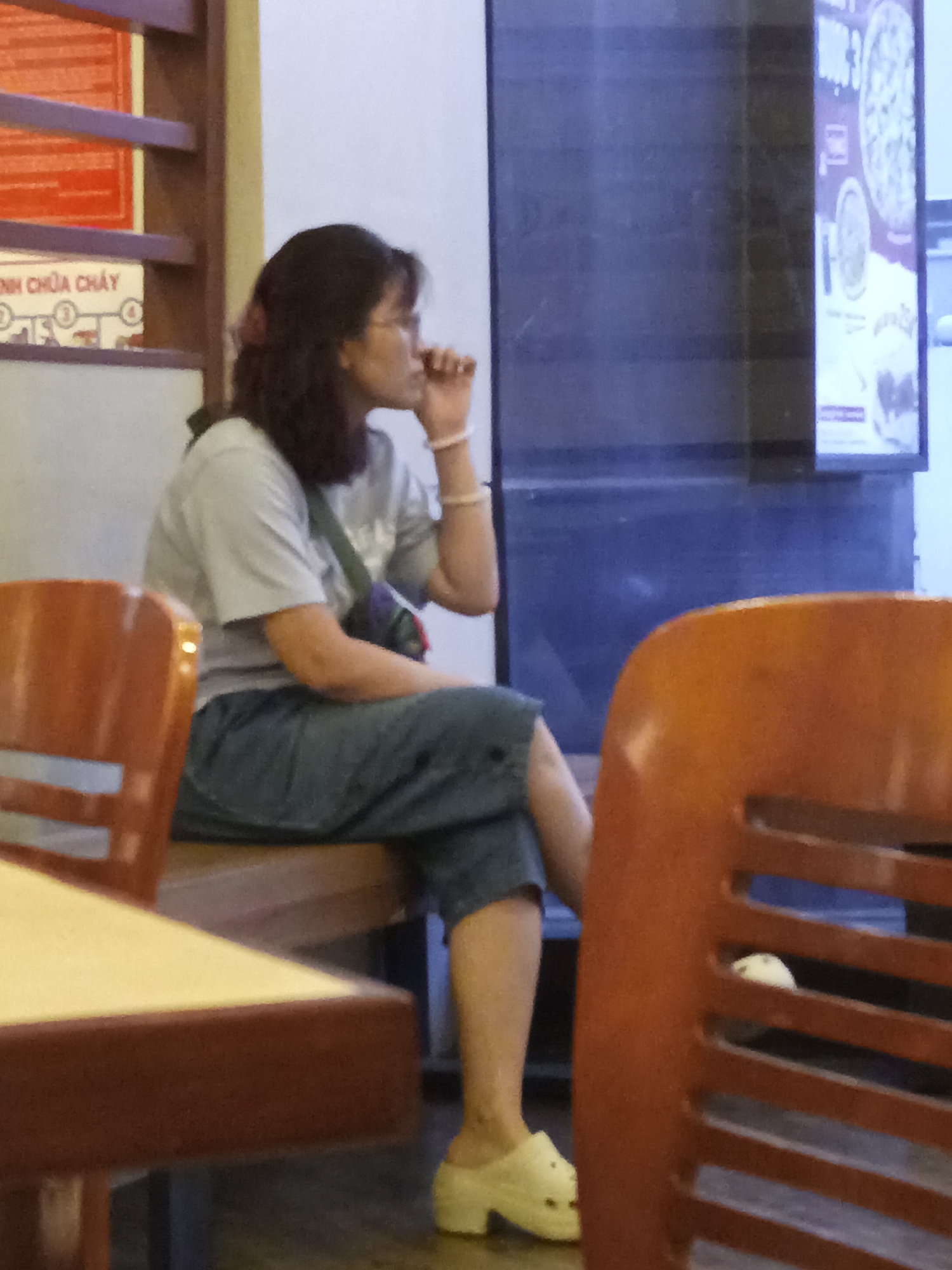 In the city of Nha Trang Ward, Vietnam, a woman of Asian descent is seated at a table, facing left. She has dark hair and is wearing glasses, a light grey t-shirt, and dark blue pants. Her right hand is near her face, touching her chin. She is wearing white slip-on shoes. A patterned bag is slung across her body. In the foreground, a dark brown wooden chair and table are partially visible. In the background, a dark blue panel and a framed menu with pizza imagery can be seen. The overall lighting is dim, with the focus on the woman.