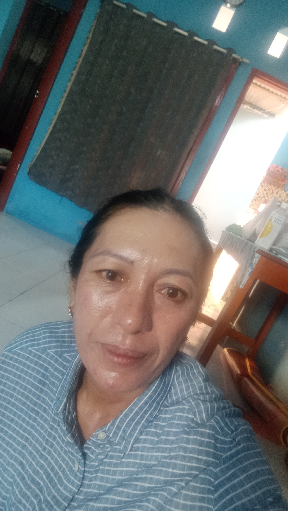 A woman is taking a selfie in what appears to be a room. She has dark hair, brown eyes, and is wearing a blue and white striped button-down shirt. Her skin appears to be dewy, suggesting it may be clean or moisturized. Behind her, the walls are painted a bright blue. To her left, there is a dark curtain hanging on the wall. To her right, there is a wooden chair with a brown seat and a reflection of what looks like a window or door. On a table next to the chair, there are some items, including a bottle with a yellow label that appears to say "LEMON WATER." The lighting suggests it may be daytime. The overall impression is an informal setting, possibly a home. The image does not contain enough contextual information to determine its specific location within Denpasar, Indonesia, beyond it being an indoor setting.