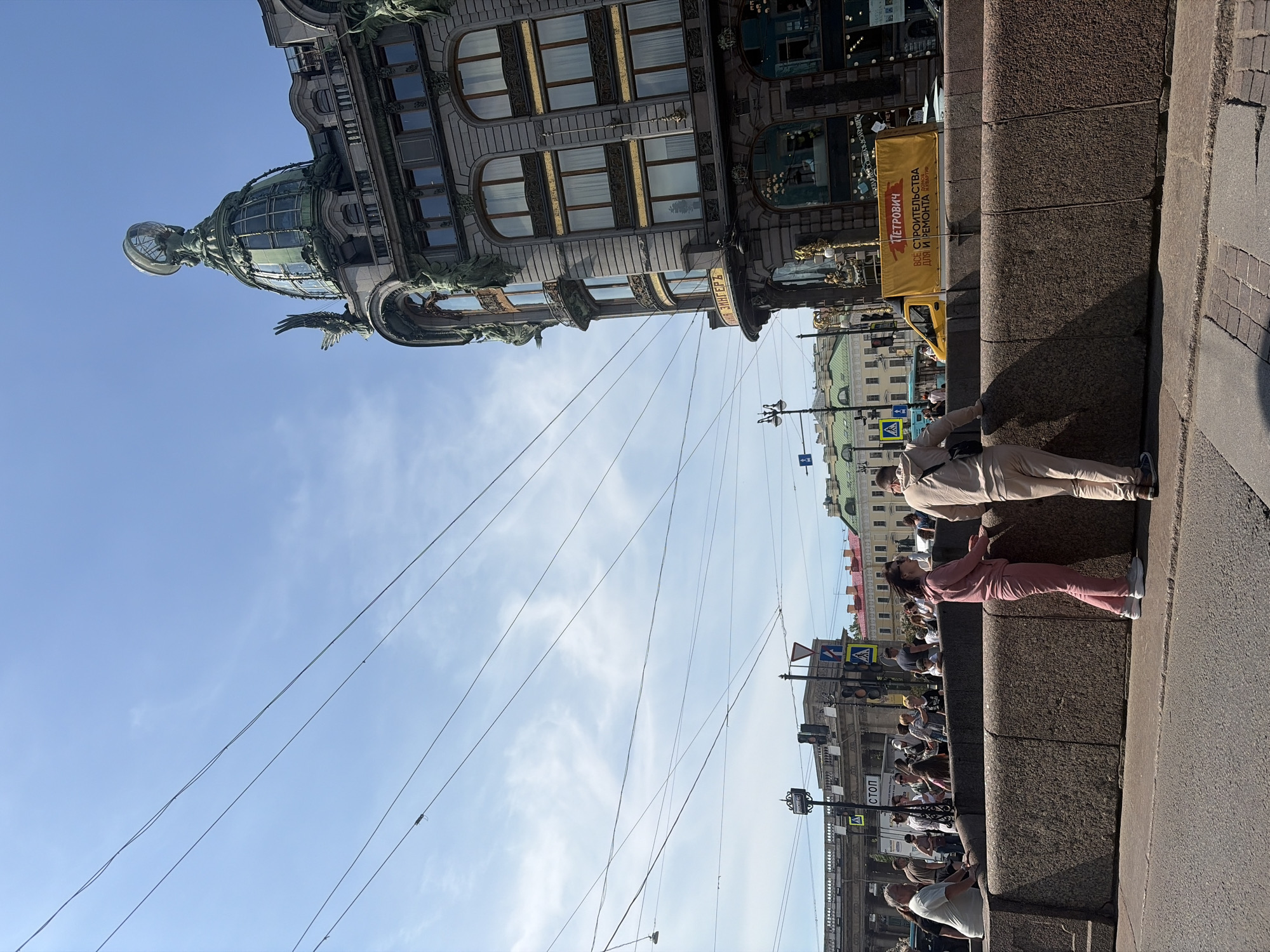 The image shows the Singer House, a prominent Art Nouveau building located at Nevsky Prospekt in Saint Petersburg, Russia. The building's iconic glass dome, topped with a globe and eagle statue, is visible against a clear blue sky with scattered clouds. In the foreground, a stone embankment runs along a street, with a few people standing on it. To the right, a yellow banner with Russian text is displayed on a vehicle. The overall scene depicts a sunny day in a bustling urban environment with a mix of architectural styles and contemporary life.