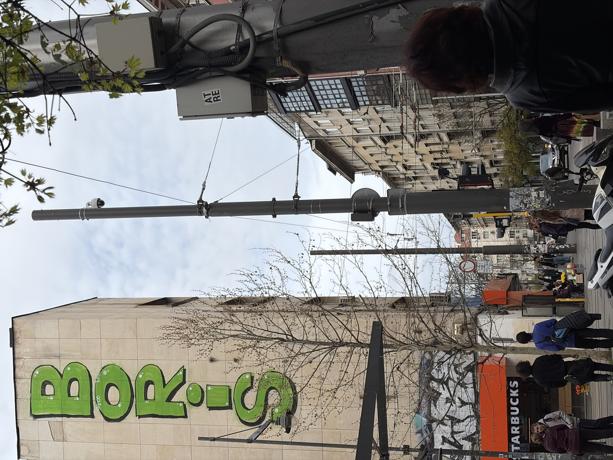 This media file captures a sideways view of an active urban street in Sofia, Bulgaria, under an overcast sky, suggesting daytime in late winter or early spring due to a prominent bare tree.
On the left side of the image (which would be the bottom if rotated correctly), a large light-colored building features bold, green graffiti spelling "BORIS". Towards the lower edge of the image (front of the street level), an orange storefront with the visible text "STARBUCKS" marks a coffee shop entrance. Several people are seen walking or standing near this area. One person wears a blue jacket and carries a bag with a "NIKE" logo.
A tall grey street pole dominates the central frame, extending vertically across the image. Attached to this pole are various wires, utility boxes, and what appears to be a CCTV camera pointed downwards. One of the utility boxes has "AT RE" printed on it. A barren tree with numerous fine branches stretches across the mid-ground.
The right side of the image (which would be the top of the street scene) reveals more multi-story buildings with numerous windows, typical of European urban architecture. Cars and a parked white scooter or motorcycle are visible on the street. A circular traffic sign, possibly indicating "no entry" or a similar restriction, is also present. People are engaged in general street activity, walking along the sidewalks.