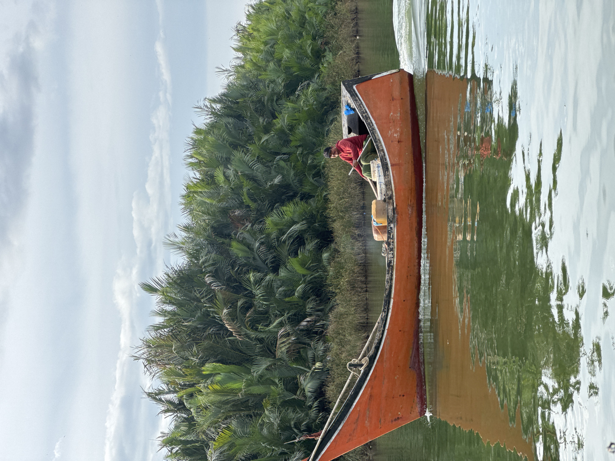 A man in a red hooded jacket and green patterned pants is rowing a narrow, orange wooden boat down a calm river. The boat has a black and white trim along its upper edge and contains two yellow containers and some blue plastic bags. The man is seated towards the stern of the boat, holding a wooden oar. The river's surface reflects the boat and the lush green vegetation on the riverbank, which is composed of dense palm-like trees with large fronds. The sky above is cloudy with patches of blue visible. The scene appears to be in a rural, natural setting, possibly in the vicinity of Mesjid Tuha, Indonesia, given the vegetation and the type of boat.