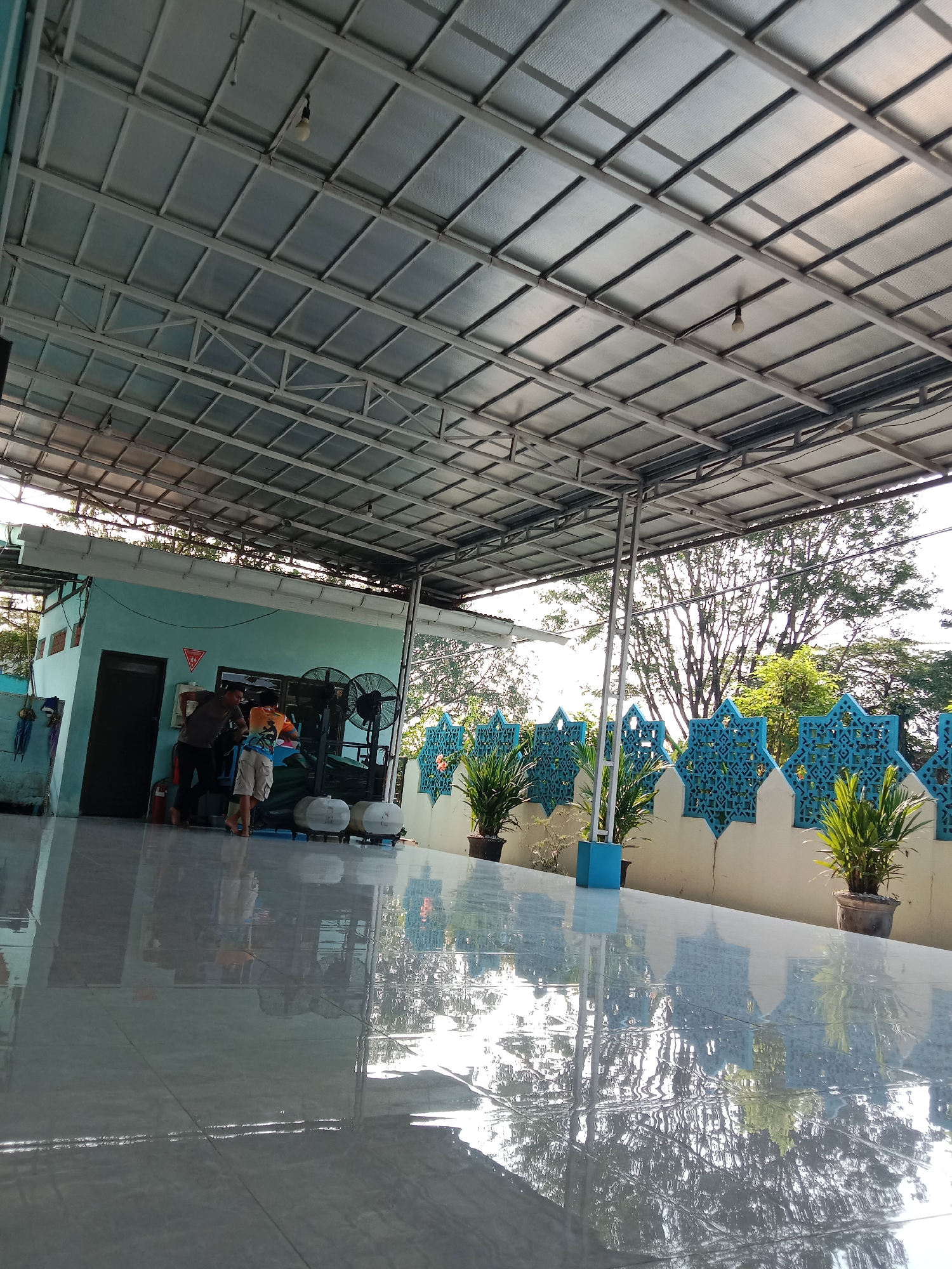 The image depicts an outdoor covered area with a metal roof supported by metal beams. The floor is tiled and highly reflective, showing mirrored images of the surroundings. In the background, a light blue building is visible, with two men standing near its entrance. One man wears a dark shirt and dark pants, while the other wears an orange shirt and light-colored shorts. Next to the building are two large fans and some white water tanks on wheels. A blue wall with decorative star-shaped cutouts runs along the right side of the scene, with potted plants placed in front of it. The overall environment appears to be an outdoor public space, possibly near a mosque or community center, given the architectural style and the presence of water tanks, which could be for ablution. The scene is likely located in the city of Semarang, Indonesia, as suggested by the visual context and common elements found in such locations in the region.