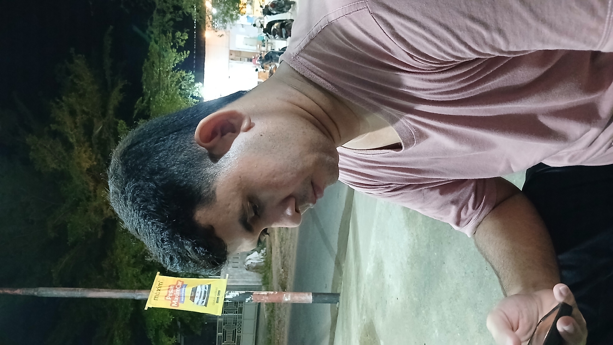 An outdoor, nighttime medium shot captures a man in Lhokseumawe, Indonesia, oriented sideways within the frame. He is seen from the chest up, with his head tilted downwards and to the right, appearing to look at something in his hand. He has short dark hair and stubble, and is wearing a light peach or pink short-sleeved t-shirt. His right arm is visible, bent at the elbow, and he is holding a dark-colored cell phone or similar device in his hand.
The background is dark, characteristic of nighttime. On the left side, lush green foliage and trees are visible, dimly lit from an unseen source. To the right and further back, there are indistinct lights, possibly from buildings or streetlights, suggesting an urban or semi-urban setting. A black pole stands upright in the middle-left of the image, to which a bright yellow banner is affixed. The banner clearly displays the text "maxim" at the top and "Pesan MaximCar" below it, indicating an advertisement for a ride-hailing or delivery service. A small image of a white car is also on the banner. The ground beneath the man appears to be paved, like a sidewalk or street. The overall atmosphere suggests a casual moment outdoors at night.
