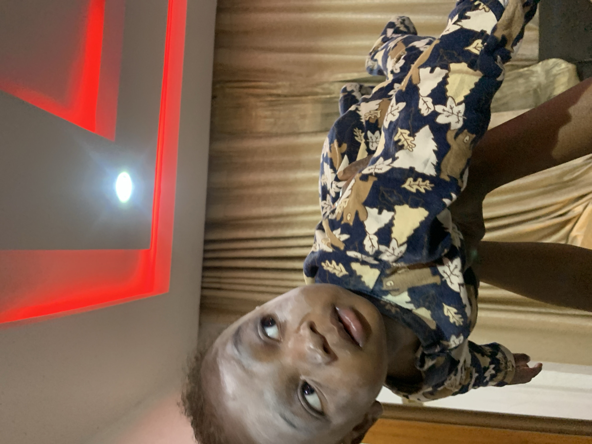 A baby is held by an adult. The baby is wearing a blue and tan leaf-patterned onesie. The baby's face is turned towards the viewer, looking up. The adult holding the baby is dark-skinned. The background is a room with a white wall that has red recessed lighting. There is a recessed light on the wall as well. The location is possibly Jos, Nigeria.
