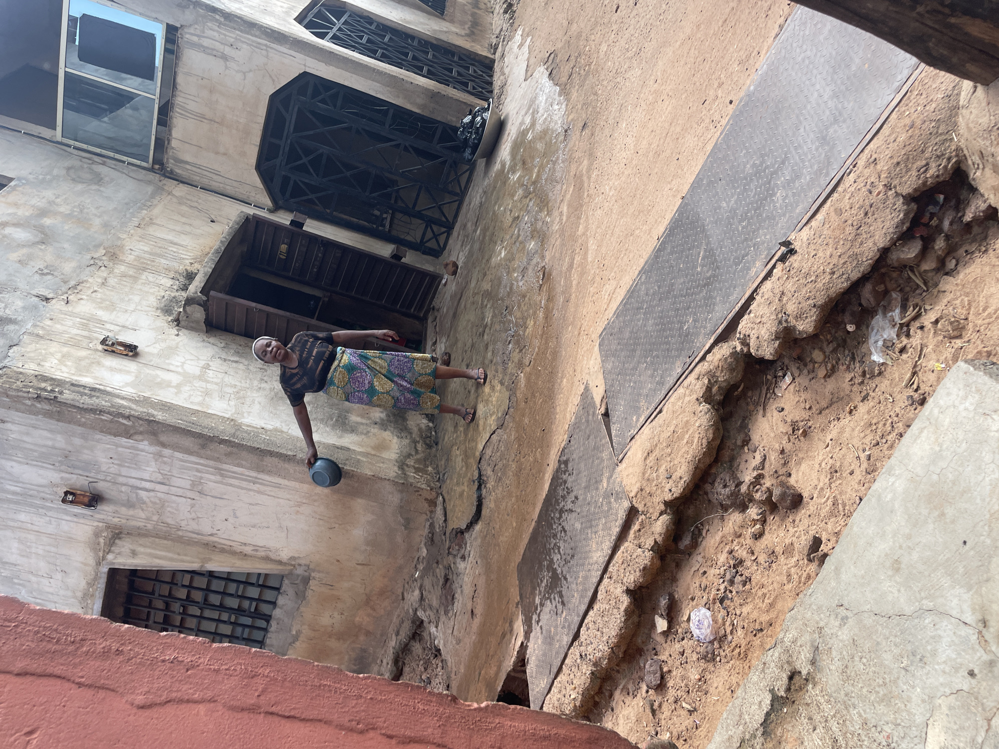 In Lokoja, Nigeria, a woman stands in a dusty, cracked courtyard in front of an old, weathered building. She is holding a small gray bowl in her right hand, which is extended slightly forward. She is wearing a dark shirt with a patterned skirt and sandals. The building has a rough, light-colored facade with several windows, some of which are barred. There are open doorways and metal security grilles visible. The ground is uneven and dirty, with patches of water and debris. A metallic ramp or cover is laid across a section of the ground in the foreground.