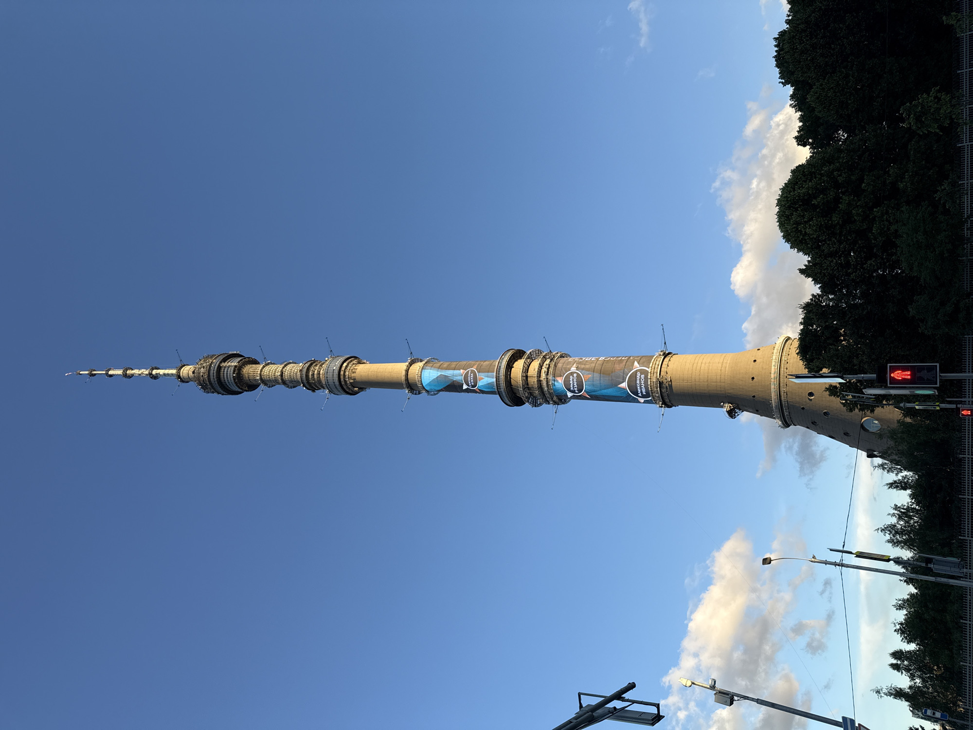 The image shows the Ostankino Tower in Moscow, Russia, under a clear blue sky with some scattered clouds. The tower, a prominent landmark, stretches upwards, its metallic structure clearly visible, featuring various platforms and antenna elements. Some circular sections of the tower are adorned with blue banners displaying white text and images, possibly related to an event or sponsorship.
In the foreground, the scene transitions to street level, with the tops of trees and a metal fence on the right. Several streetlights and traffic signals are visible, including a red pedestrian crossing signal indicating it's unsafe to cross. The overall atmosphere suggests it is daytime, likely late afternoon or early evening given the quality of light. The setting is urban, with infrastructure typical of a city environment. There are no people visible in the frame, and the focus is on the architecture of the tower and the surrounding urban landscape.