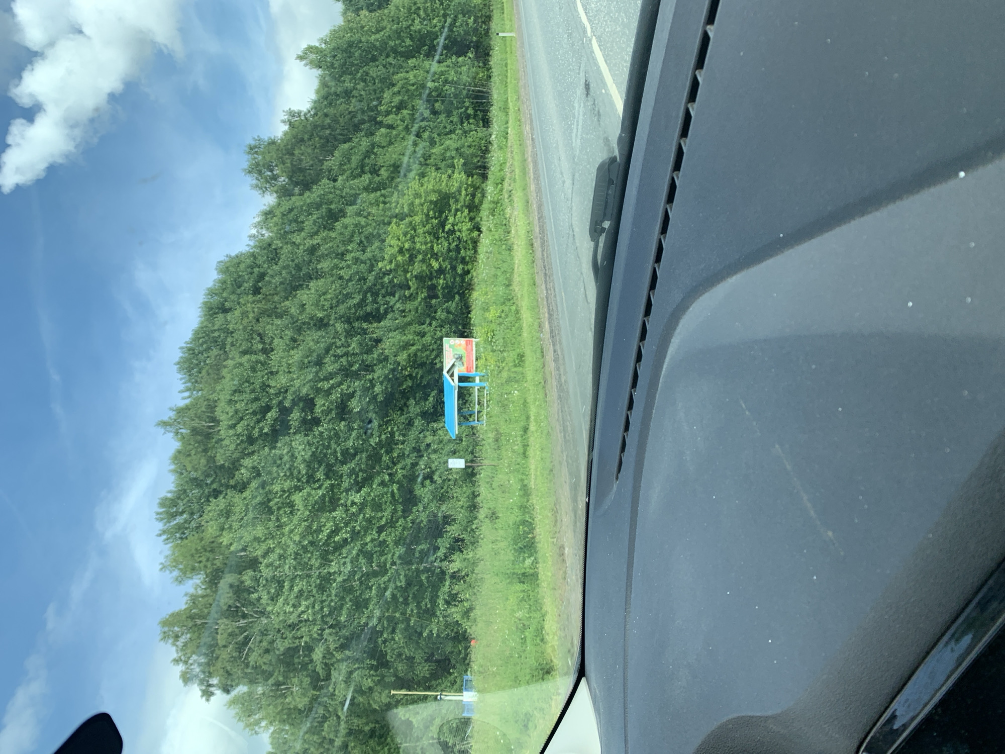 This image, taken from inside a vehicle, captures a roadside scene on a bright, cloudy day. The foreground shows the dark dashboard of the car and a portion of the windshield. Beyond the windshield, a paved road stretches into the distance. To the left of the road, a grassy verge is bordered by a dense line of green trees forming a backdrop. In the middle ground, a small, brightly colored structure resembling a bus stop shelter with a blue roof and red and white walls is visible. A white signpost stands to its right. Further to the left, a yellow post and a white structure are partially obscured by the trees. The sky above is a bright blue with scattered white clouds. The location is identified as being in the city of Sudak, Russia.