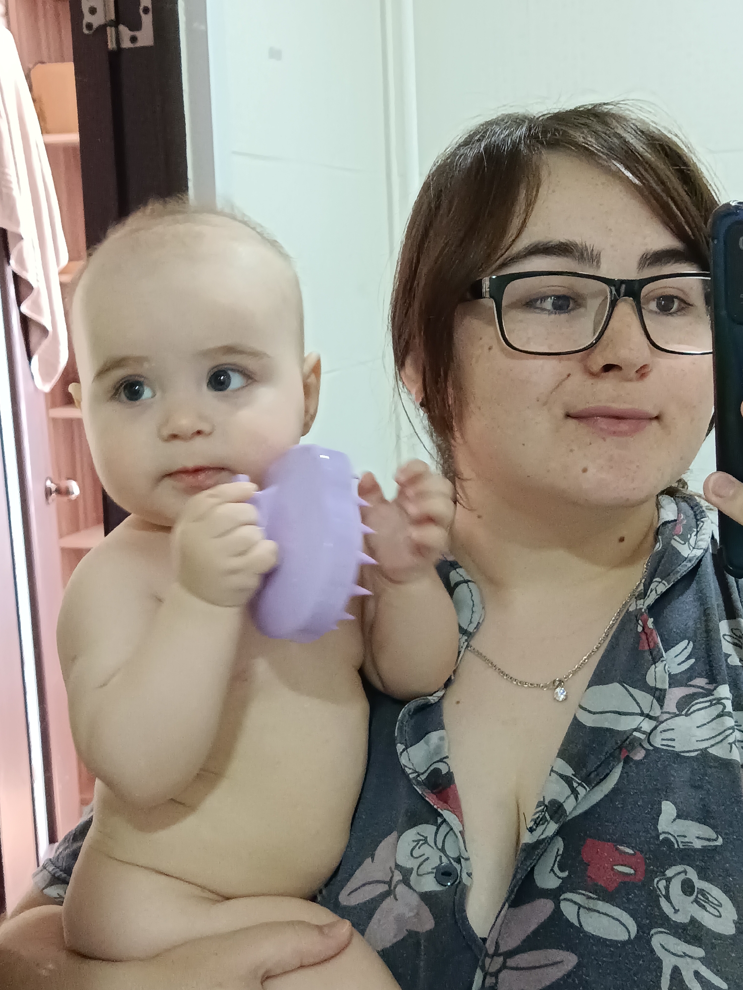 A woman with short brown hair and glasses holds a baby. The baby is holding a purple, spiky toy. The woman is wearing a navy blue shirt with a Mickey Mouse pattern and a silver necklace. The scene appears to be in a bathroom. The city and location are unknown.