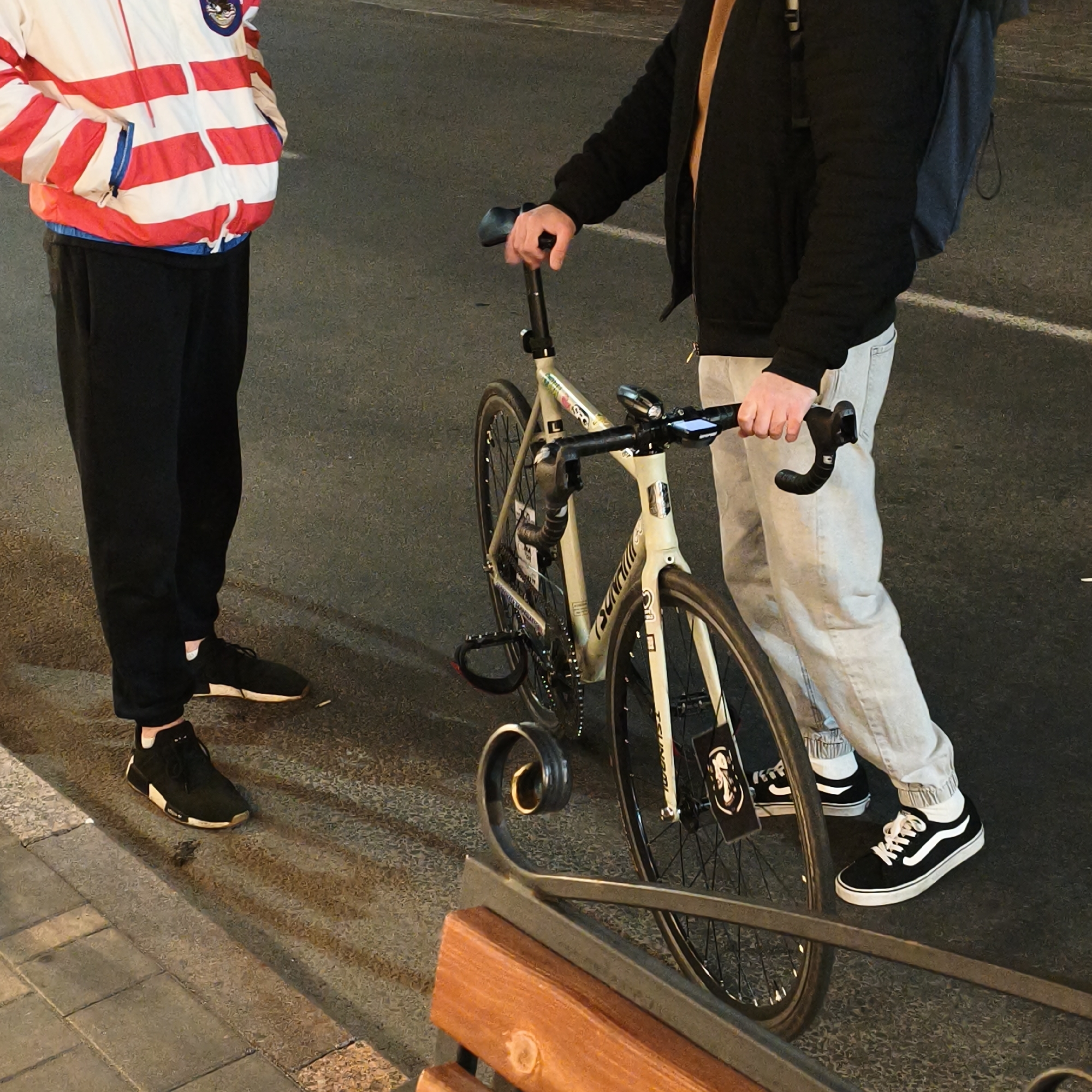 The image shows two people standing near a bicycle and a bench. One person is wearing a red and white striped jacket, black pants, and black sneakers with white accents. The other is wearing a black jacket, tan shirt, grey jeans, and black and white sneakers. The bicycle is a light-colored road bike with black tires and a front light. The bench is made of wood and metal. The location appears to be a street in Hrodna, Belarus.