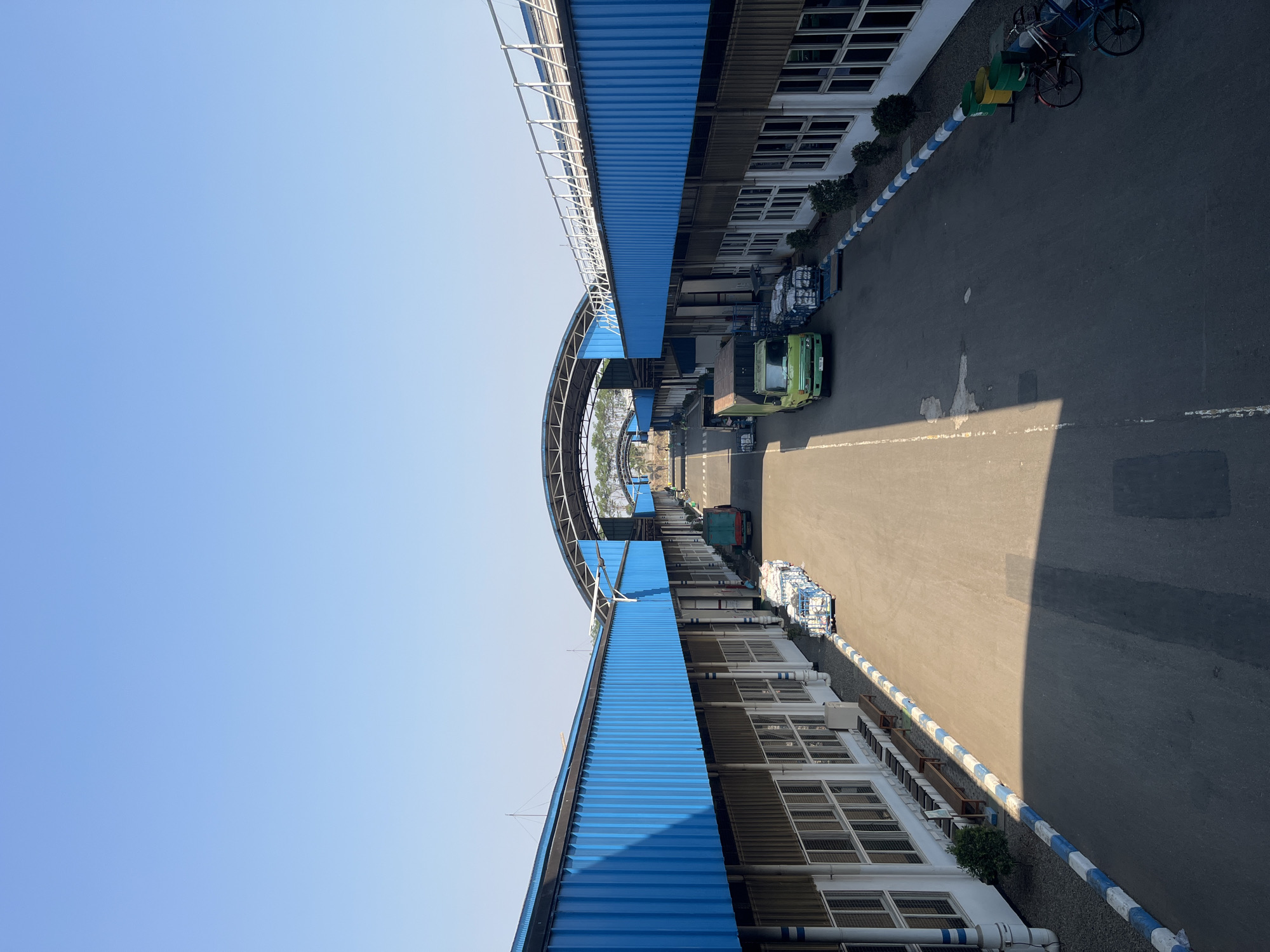 The image depicts an industrial area, likely a warehouse or factory, with a clear blue sky overhead. The architecture features long, low-slung buildings with bright blue corrugated metal roofs and white walls. The buildings are arranged symmetrically, creating a pathway between them that leads into the distance.
Along the pathway, there are several vehicles, including a green truck and smaller utility vehicles. Carts filled with what appear to be packaged goods are also visible. The ground is paved asphalt.
On the left side of the pathway, in front of the building, there are wooden benches and some greenery. On the right side, there are rows of windows, more greenery, trash cans, and bicycles parked near the edge. The scene appears to be quiet and operational, with no people prominently featured in action. The lighting suggests it is daytime, likely morning or afternoon, with strong shadows cast on the ground. The location is identified as Pelang, Indonesia, suggesting a tropical or subtropical climate based on the clear sky and vegetation.