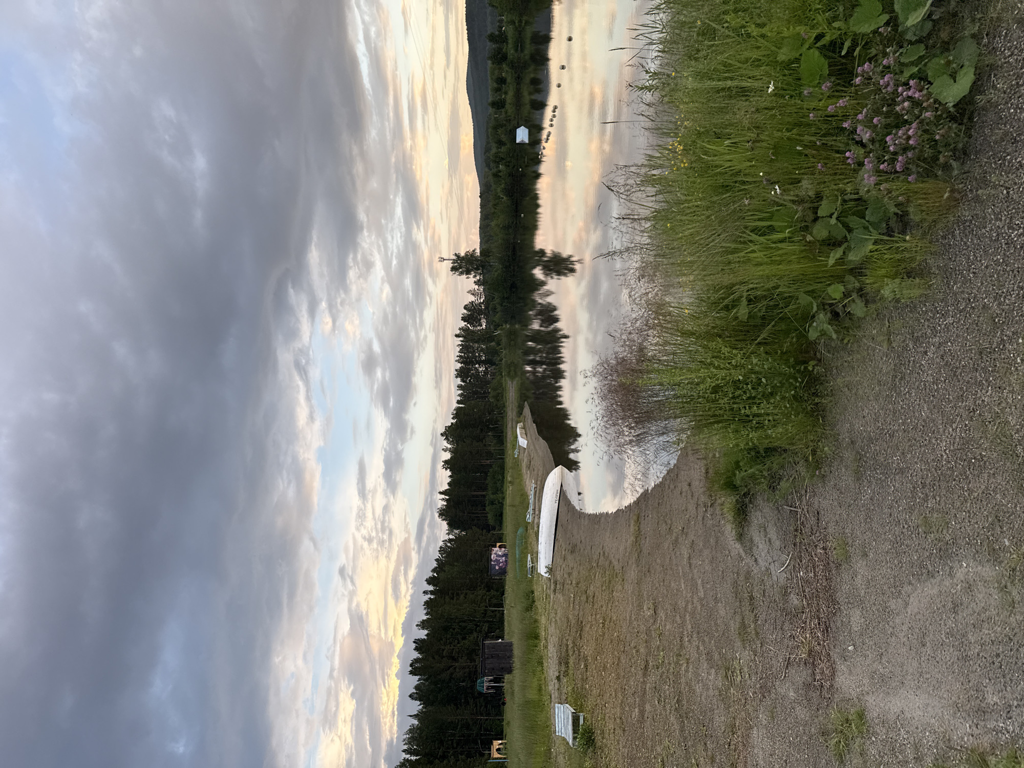 The image depicts a serene lakeside scene at dusk in Syrostan, Russia. A calm body of water reflects the soft hues of a cloudy sky, with the setting sun casting a warm glow. The shoreline is a mix of sand and gravel, with a patch of lush green grass and wildflowers in the foreground. Several upturned boats are visible on the beach, suggesting a recreational area. In the background, a dense forest lines the opposite bank of the lake, with a few small structures and a communication tower dotting the landscape. The overall atmosphere is peaceful and tranquil, characteristic of a quiet evening by the water.