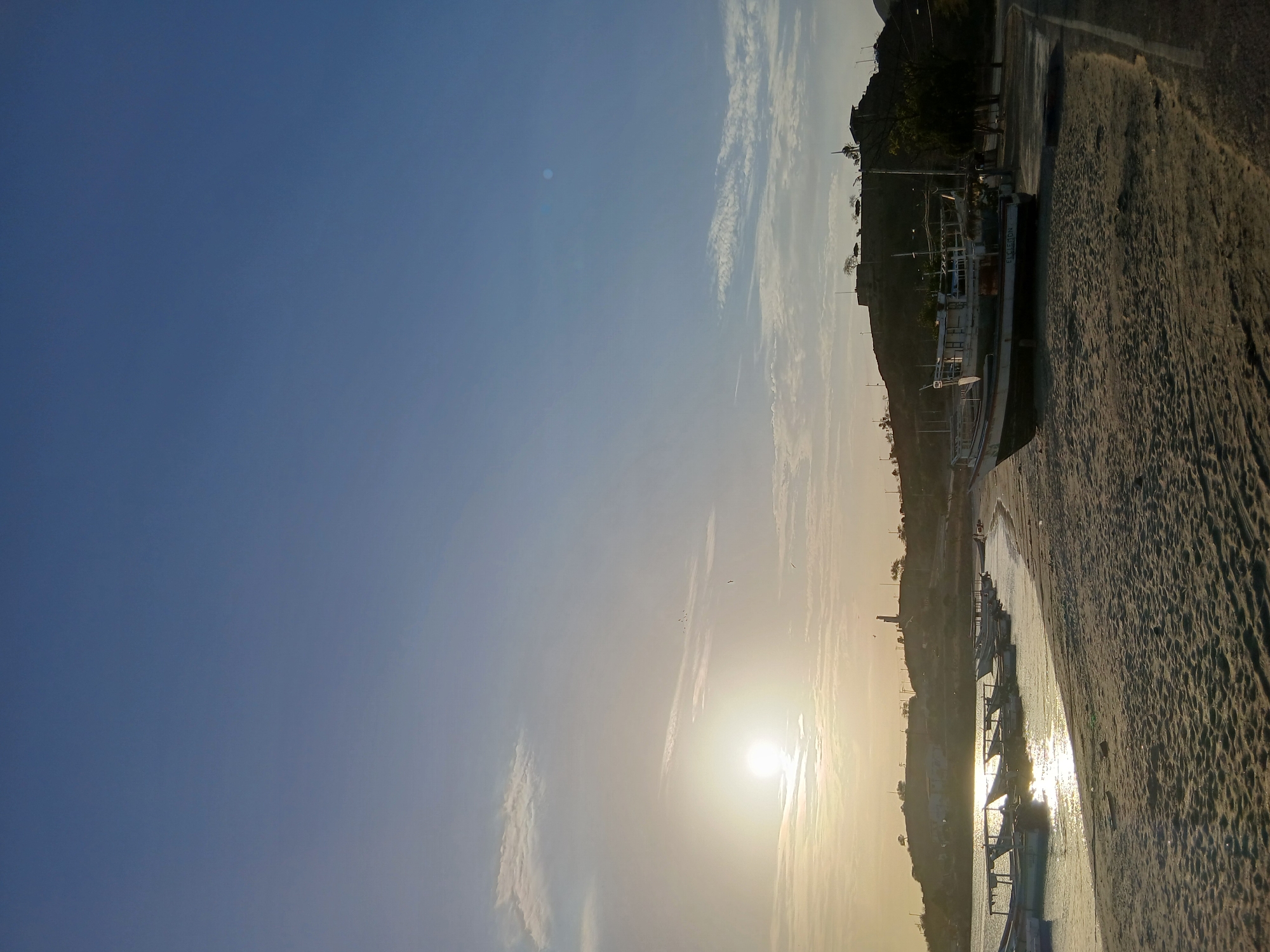 The image shows a sunset in Juan Griego, Venezuela. The sun is low in the sky, casting a bright glare on the water and highlighting the sandy beach. Several fishing boats are anchored in the shallow water, their silhouettes visible against the sun's reflection. On the shore, a large concrete structure is observed, which is one of the fishing boats. The sky transitions from a deep blue at the top to a hazy yellow-orange near the horizon.