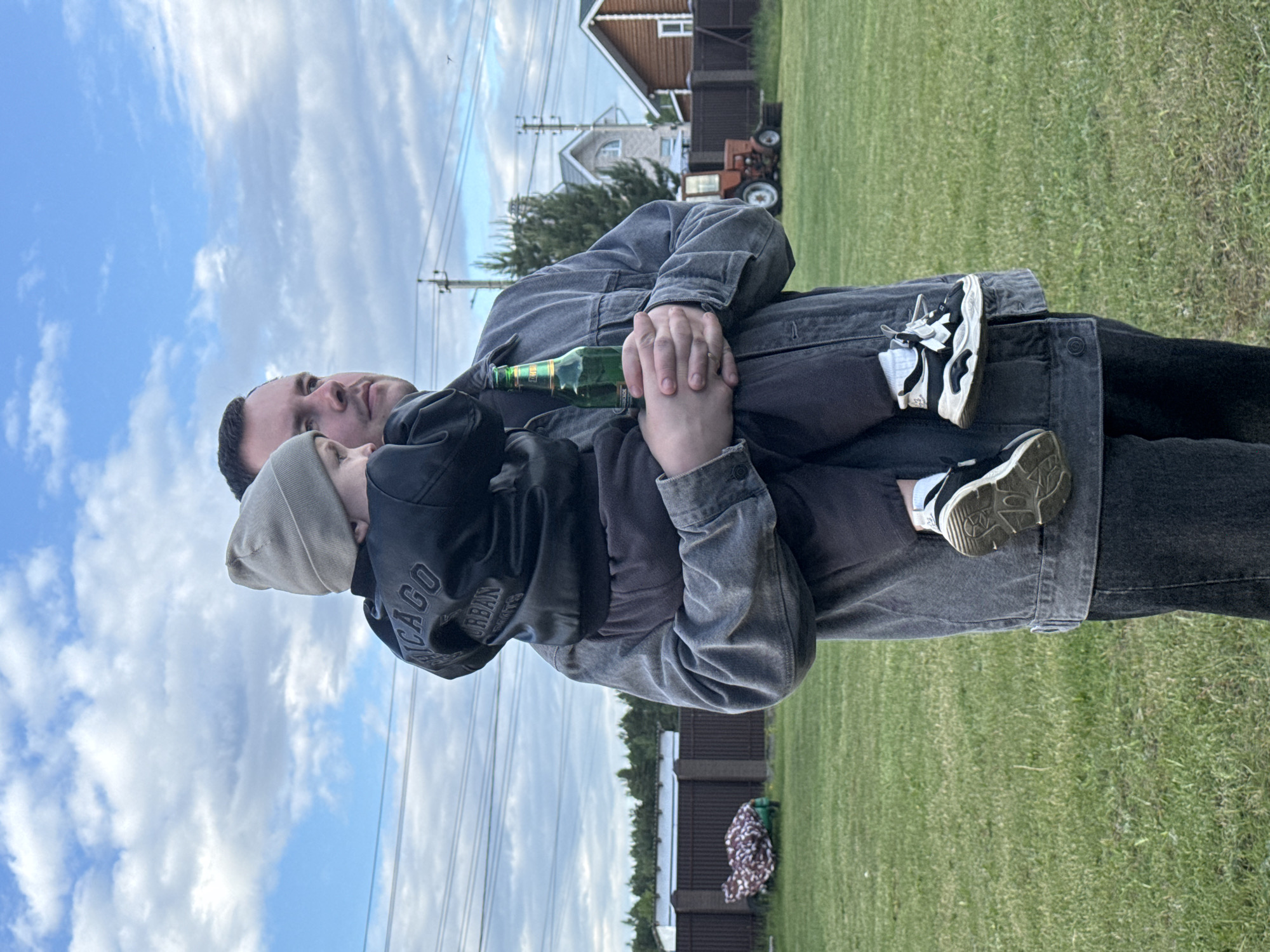 A man holds a child, both facing upwards towards the sky. The man wears a grey jacket and dark pants. He holds a green bottle in his hands. The child wears a dark hoodie with "Chicago Urban" visible on the sleeve and a light-colored beanie. They are standing on a grassy lawn. In the background is a house, trees, a fence, and a utility pole with wires. An orange tractor is also visible. The location appears to be in the city of Vysokinichi, Russia.