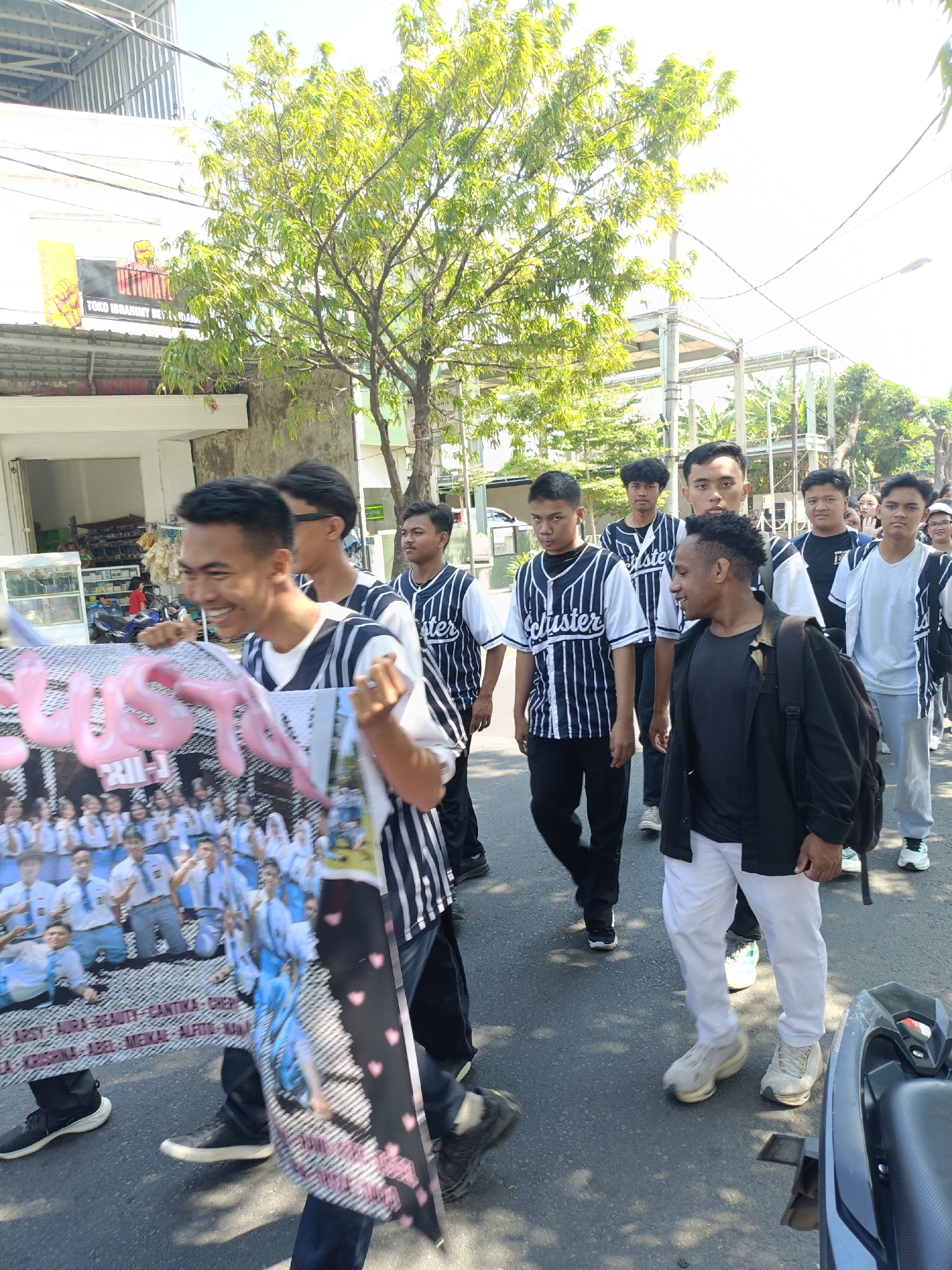 A group of young men, some wearing matching striped baseball jerseys, walk down a street in Kediri City, Indonesia. The man in the foreground, holding a banner with pink lettering and photos of people in school uniforms, smiles broadly. The banner displays names like "ARSY," "AURA," "BEAUTY," and "CHANTIKA." Behind him, other young men are also walking, some looking towards the camera, others straight ahead. The street appears to be lined with shops and trees, with a bright, sunny sky overhead. A parked motorcycle is visible on the right side of the image. The overall atmosphere suggests a celebration or a parade, possibly related to a school or graduation event.
