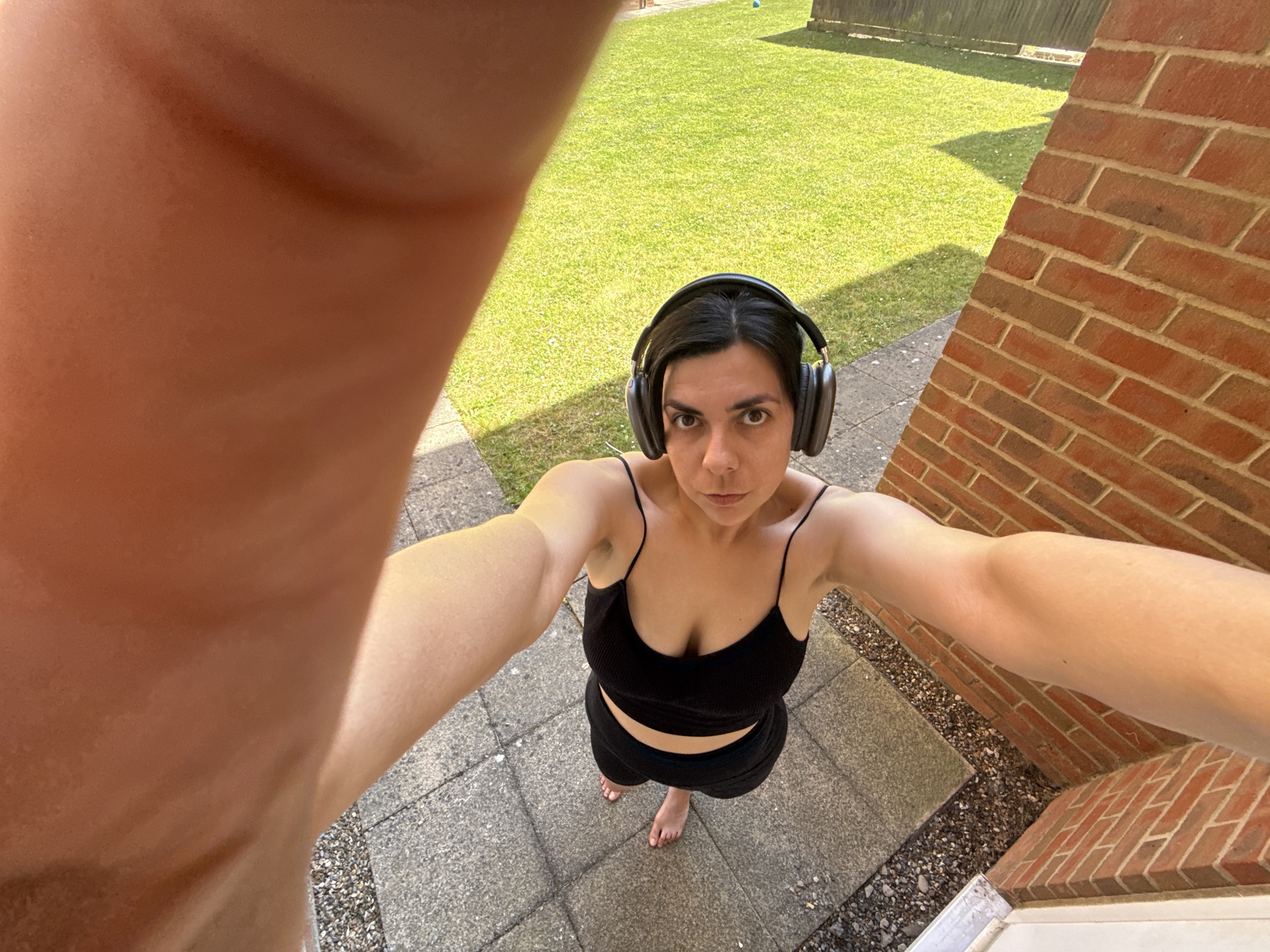 A high-angle selfie photograph shows a woman standing outdoors on a paved area next to a brick wall and a grassy yard. The woman is wearing a black tank top and shorts, and she has headphones on. Her arms are outstretched toward the camera, causing the foreground to be dominated by a blurry view of her arm. Behind her, there is a green lawn that is bordered by a wooden fence. The location appears to be outdoors in a residential area in the city of Great Yarmouth in the United Kingdom.