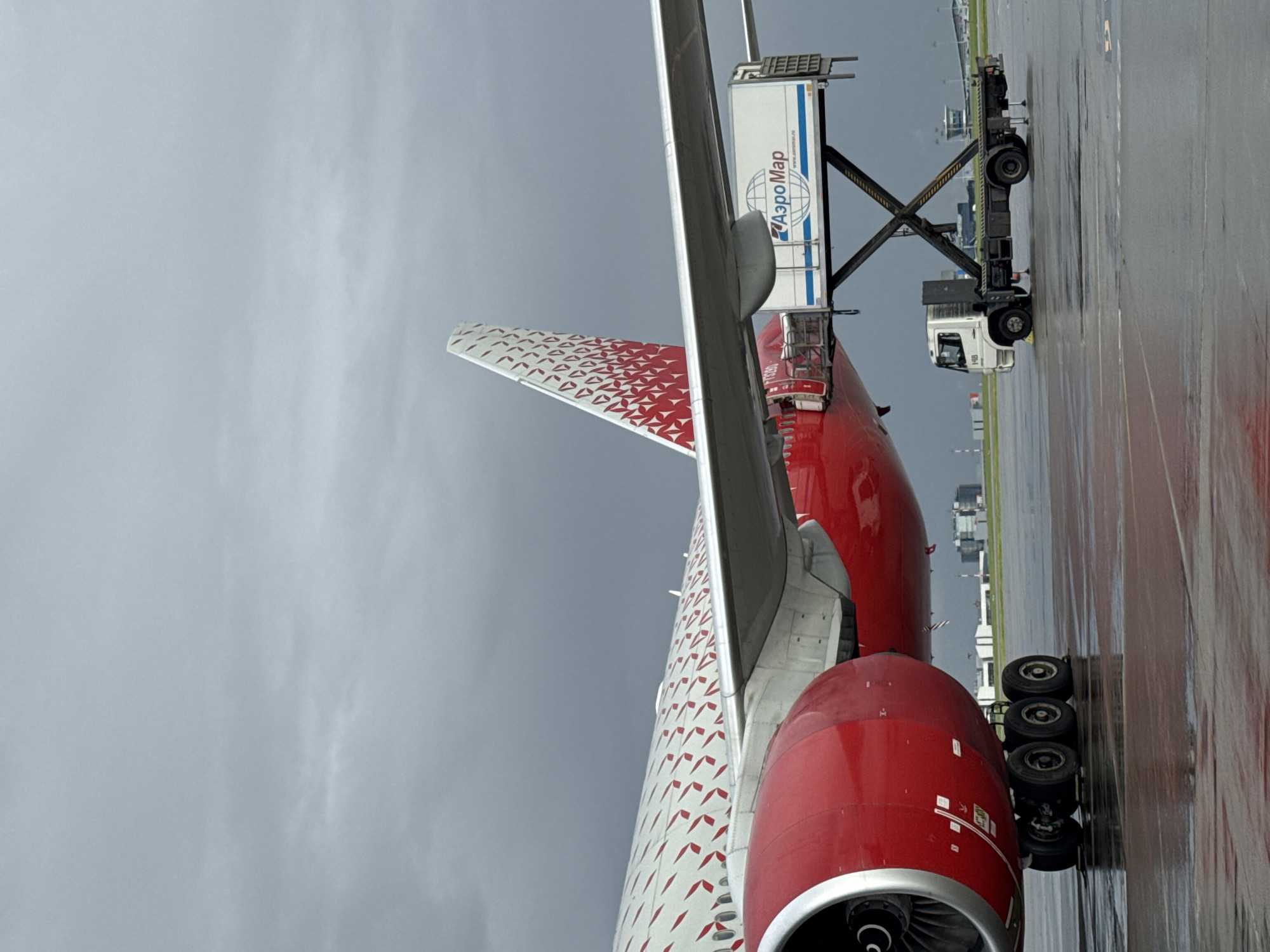 The image shows a close-up, low-angle view of the rear section of a red and white passenger airplane on a wet tarmac. The airplane's tail fin, partially visible, is white with a repeating pattern of small red shapes. The main body of the aircraft is red, and a large red engine nacelle with a visible turbine is in the foreground. The landing gear, consisting of a set of three wheels, is also visible.
A baggage loader vehicle is positioned adjacent to the aircraft, with its platform extended. The side of the loader's container has text that reads "АэроМар" (Aeromar) in blue Cyrillic letters, with a globe logo. Below this, smaller text is visible, including "www.aeromar.ru". A white cab with the number "301" is visible on the loader vehicle.
The scene is set at an airport, with other airport buildings and infrastructure faintly visible in the background. The sky is overcast and gray, suggesting a cloudy or rainy day. The tarmac is wet and reflective, indicating recent rain. The overall atmosphere is utilitarian, focused on aircraft operations. No people are clearly visible. The image is oriented vertically.