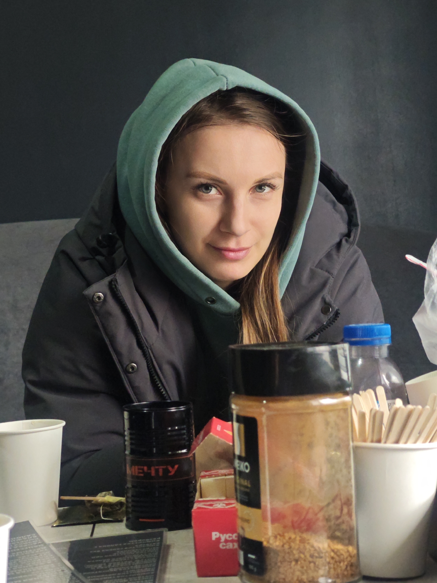 A medium shot of a young, light-skinned woman with green eyes and light brown hair, wearing a light green hooded sweatshirt under a dark gray puffy jacket, sitting at a table cluttered with various items. In front of her, on the table, are two white paper cups, a black cylindrical can with the word "МЕЧТУ" in red lettering, a red box, a clear plastic bottle with a blue cap, and a clear plastic spice jar with a black cap. The background is a solid gray wall, and the location is Unknown, Unknown.