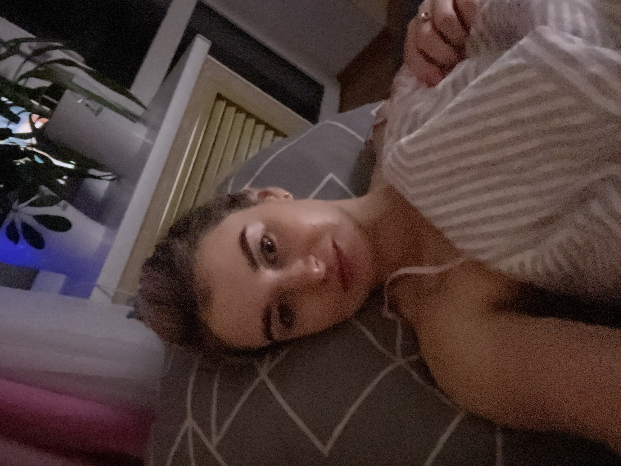 A woman lies on a chevron-patterned pillow, partially covered by a striped garment. Her head rests on the pillow, her face tilted towards the camera. A potted plant with large, dark leaves is visible in the background near a window with dark surroundings suggesting night. The interior appears to be a residential setting. The specific location in Chaikovsky, Russia, is not discernible from the image.