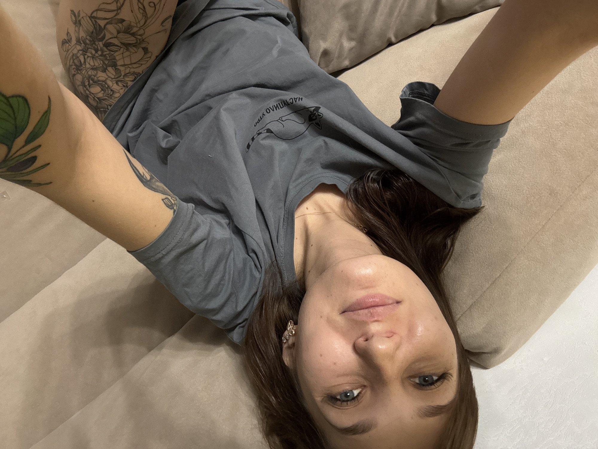 A young woman is lying on a sofa, taking a selfie. She is wearing a gray t-shirt with a graphic of a raccoon and Russian text that reads "Наступило утро" (It's morning). Her right arm is visible, adorned with tattoos of flowers and a bird. The sofa is a light beige color. The background is a white textured wall. The image was taken indoors, likely in a residential setting in Novokuznetsk, Russia.