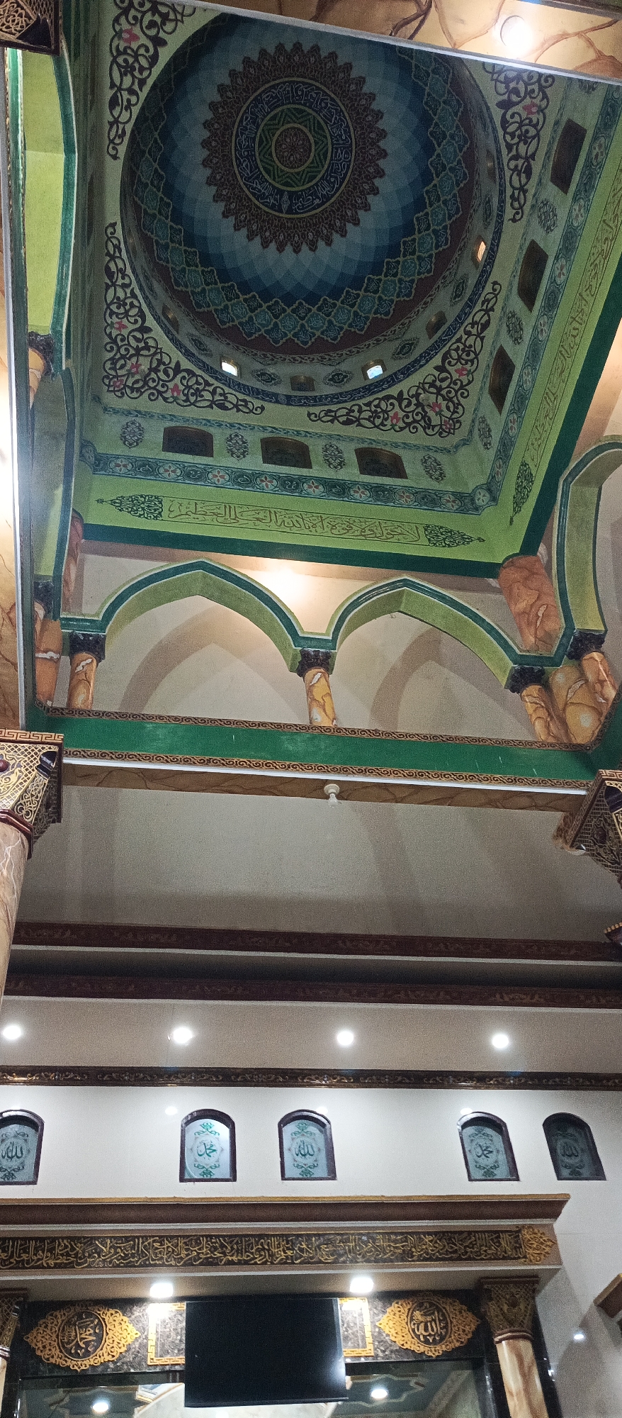 The image captures an interior view of a mosque, likely within the city of Arjasa, Indonesia. The architectural style is evident in the ornate ceiling, featuring a large, intricately decorated dome with Islamic calligraphy and geometric patterns. The dome is painted in shades of blue, green, and brown, with a central circular motif.
Below the dome, the ceiling transitions to a green panel adorned with more calligraphy and decorative elements. This green band extends outwards, meeting supporting structures that resemble arches. These arches are supported by decorative pillars, some of which have a marble-like finish in brown and beige.
Further down, the scene reveals a wall with several arched niches. Three of these niches are clearly visible, each containing an Islamic inscription within a green frame. The wall itself is a light cream color, with decorative borders above and below the niches.
At the bottom of the image, a horizontal decorative element in gold and dark brown spans the width. A black rectangular screen, possibly a monitor or display, is partially visible in the lower center, flanked by ornate gold frames containing Islamic calligraphy. The overall impression is one of rich Islamic artistry and religious significance.
