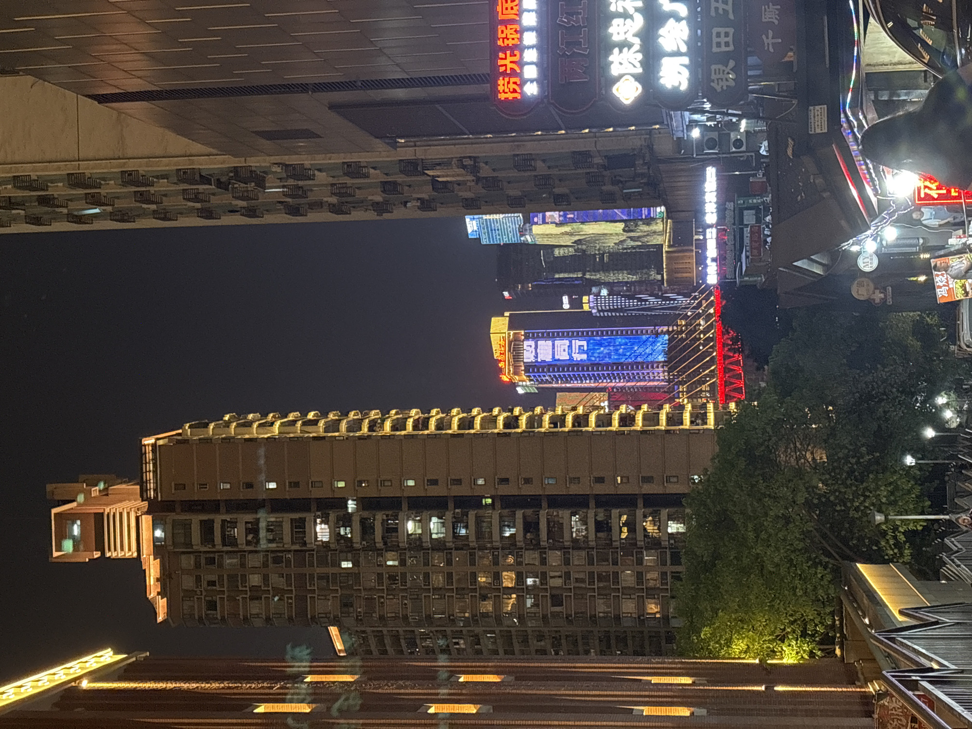 This is a nighttime view in the Yuzhong District of China, showcasing a cluster of illuminated buildings. The buildings feature windows with visible lights and are adorned with various signs in Chinese characters. Streetlights are scattered around, contributing to the overall brightness of the scene. The setting is urban, emphasizing the blend of modern architecture and traditional signage within the bustling city.