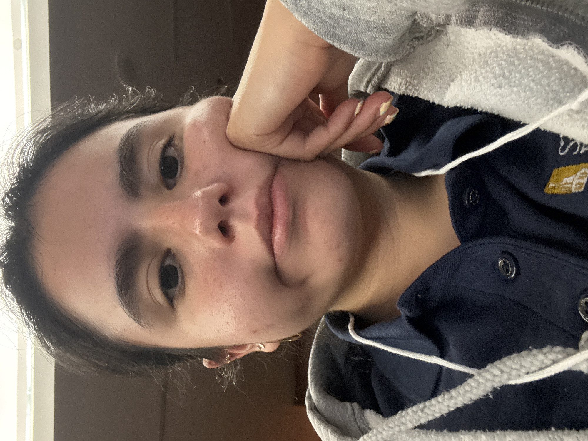 A close-up, slightly off-center portrait of a young woman with dark hair. Her left hand is gently resting on her cheek. She is wearing a dark blue collared shirt, which appears to be a school uniform or casual jacket, with a visible white drawstring. Over the blue garment is a gray hooded sweatshirt. The background is out of focus. No information in the image indicates a location in San Antonio, United States of America.